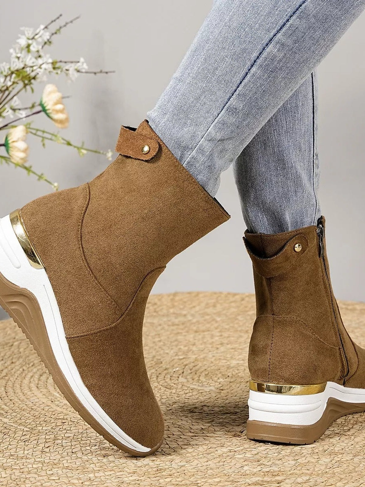 A model wears brown suede ankle boots with wedge heels, gold-tone toe caps and trim, side zippers, and decorative ankle straps over light jeans.