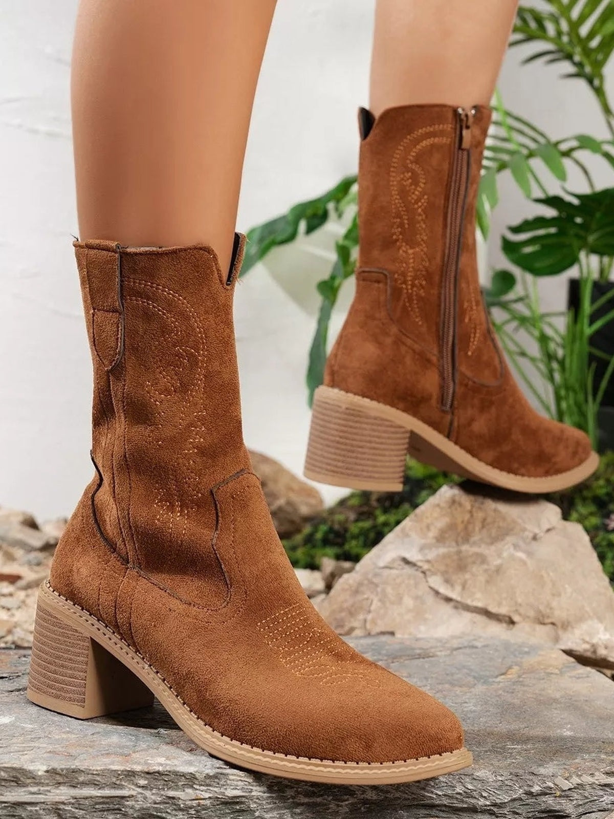Model wears brown suede-look Western boots with mid block heels, pointed toes, decorative stitched panels, and side zippers in a staggered pose.