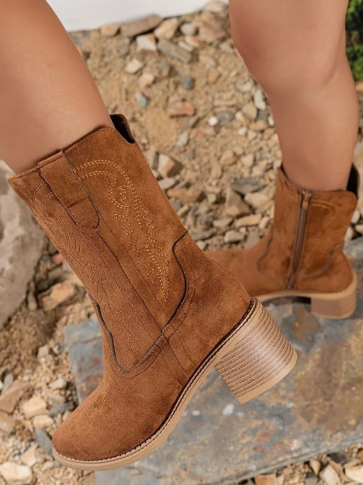 A model wears brown suede western boots with stitched shaft detailing, rounded toes, side pull tabs, and mid-height block heels.