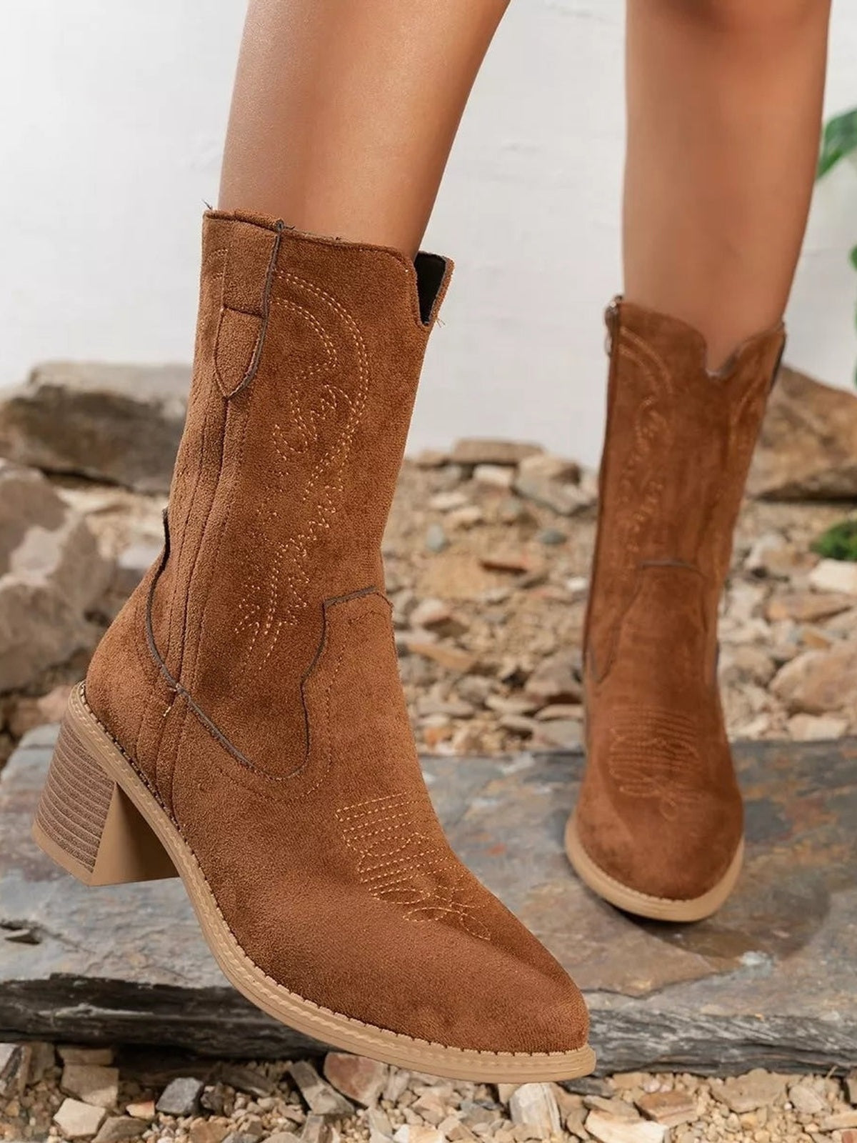 Brown suede-style western boots with pointed toes, decorative stitching, tall shafts, and stacked block heels, shown worn by a model.