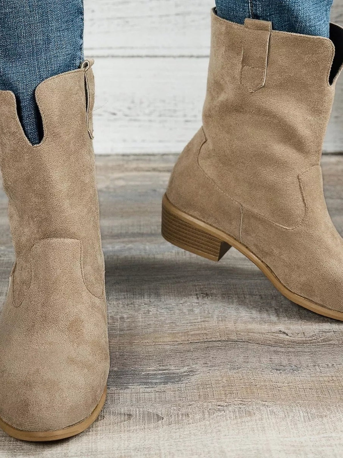 Tan suede western boots on a model in blue jeans, featuring pull tabs, stitched panel construction, rounded toes, and stacked mid-height block heels.