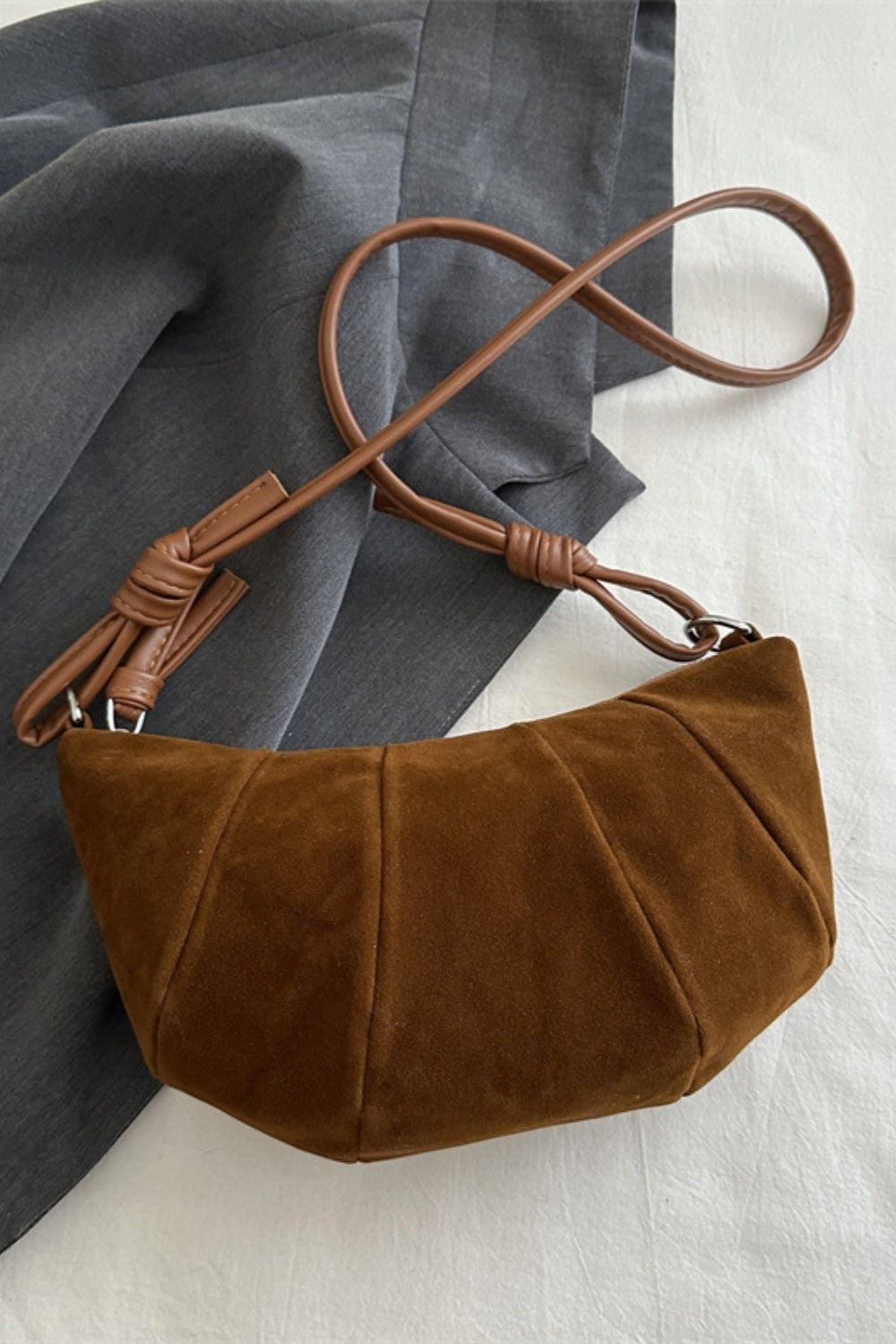 Brown suede croissant-shaped shoulder bag with a softly curved silhouette, gathered paneled construction, and a long adjustable strap.