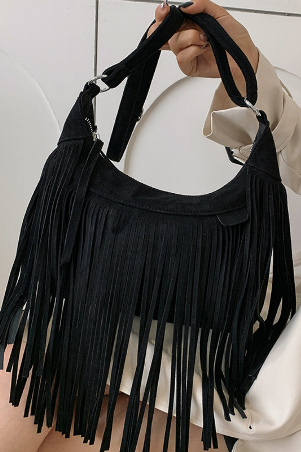 A model holds a black suede fringe shoulder bag with a softly curved silhouette, long fluid fringe, silver hardware, and an adjustable strap.