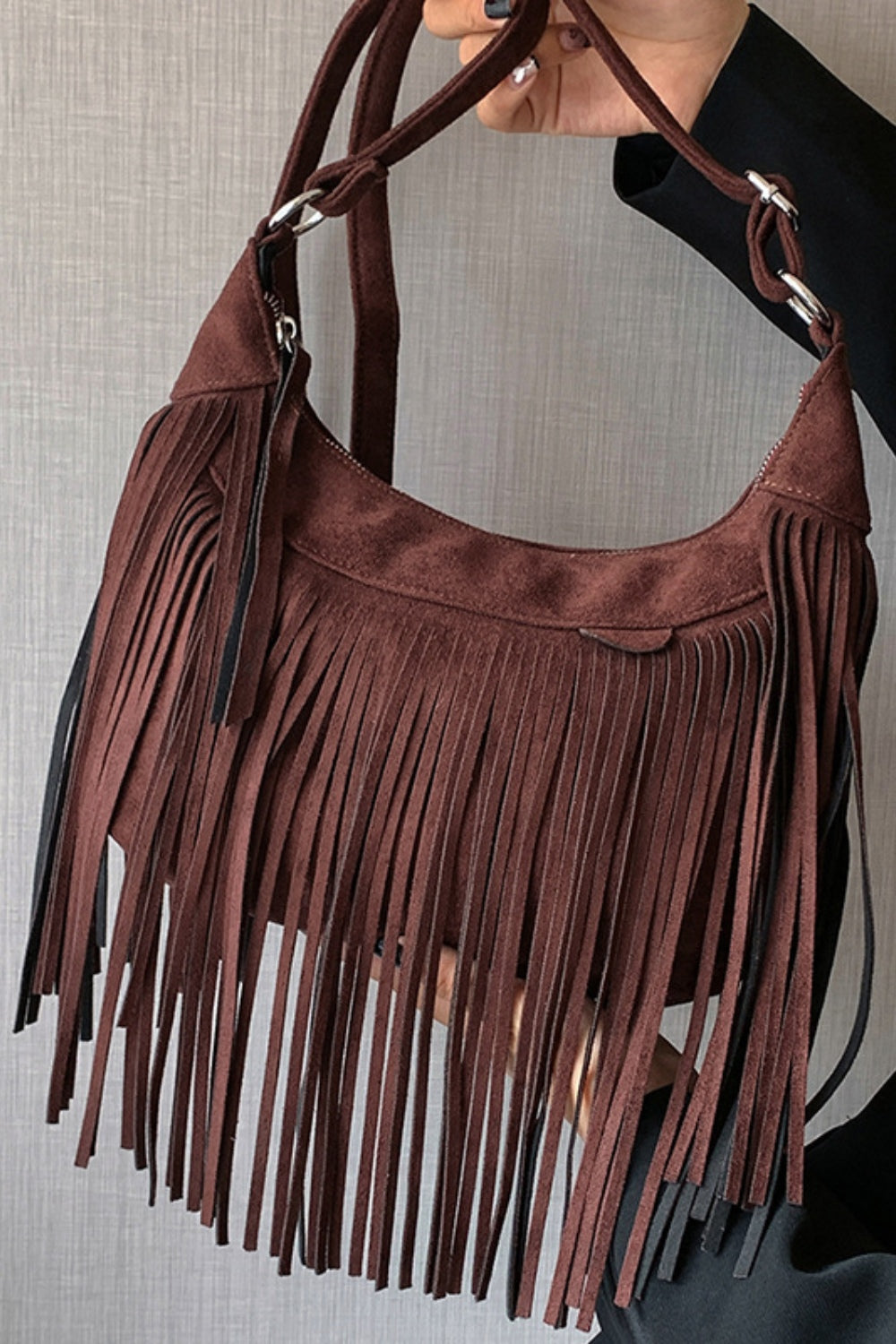 A close-up shows a brown suede fringe shoulder bag with a softly curved silhouette, long layered fringe, and adjustable strap held by a model.