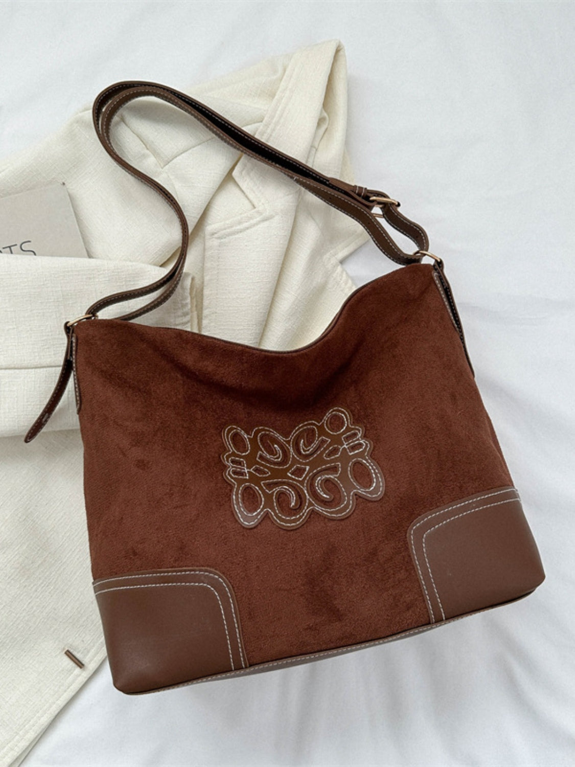 Medium brown suede patch tote bag with a relaxed silhouette, curved upper edge, adjustable PU leather strap, stitched front motif, and structured lower panels.