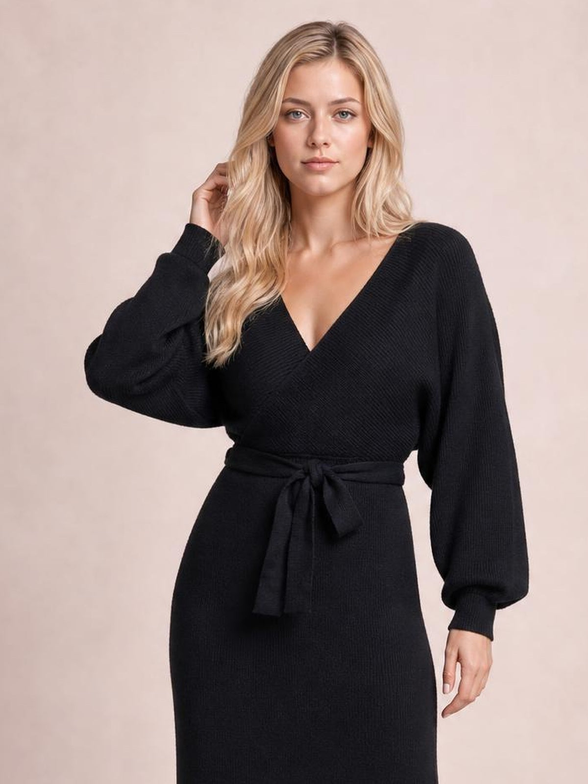 Model poses in a black sweater dress with a surplice neckline, long sleeves, fitted silhouette, and tied bow waist detail.