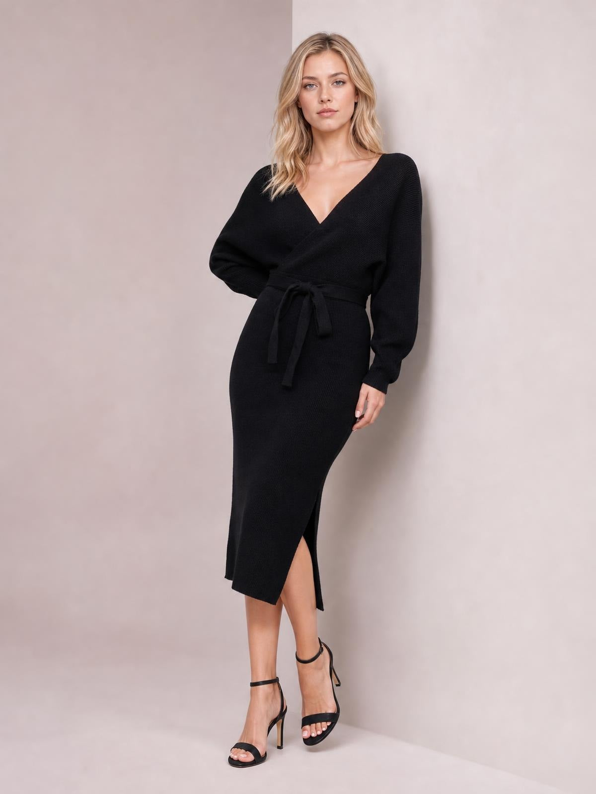 A model poses in a black surplice-neck sweater dress with long sleeves, a tied bow waist, fitted midi silhouette, and side slit.