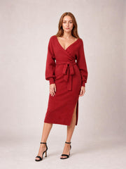 Model in a red surplice-neck sweater dress with long sleeves, a tied bow waist, fitted midi silhouette, and side slit, standing in heels.