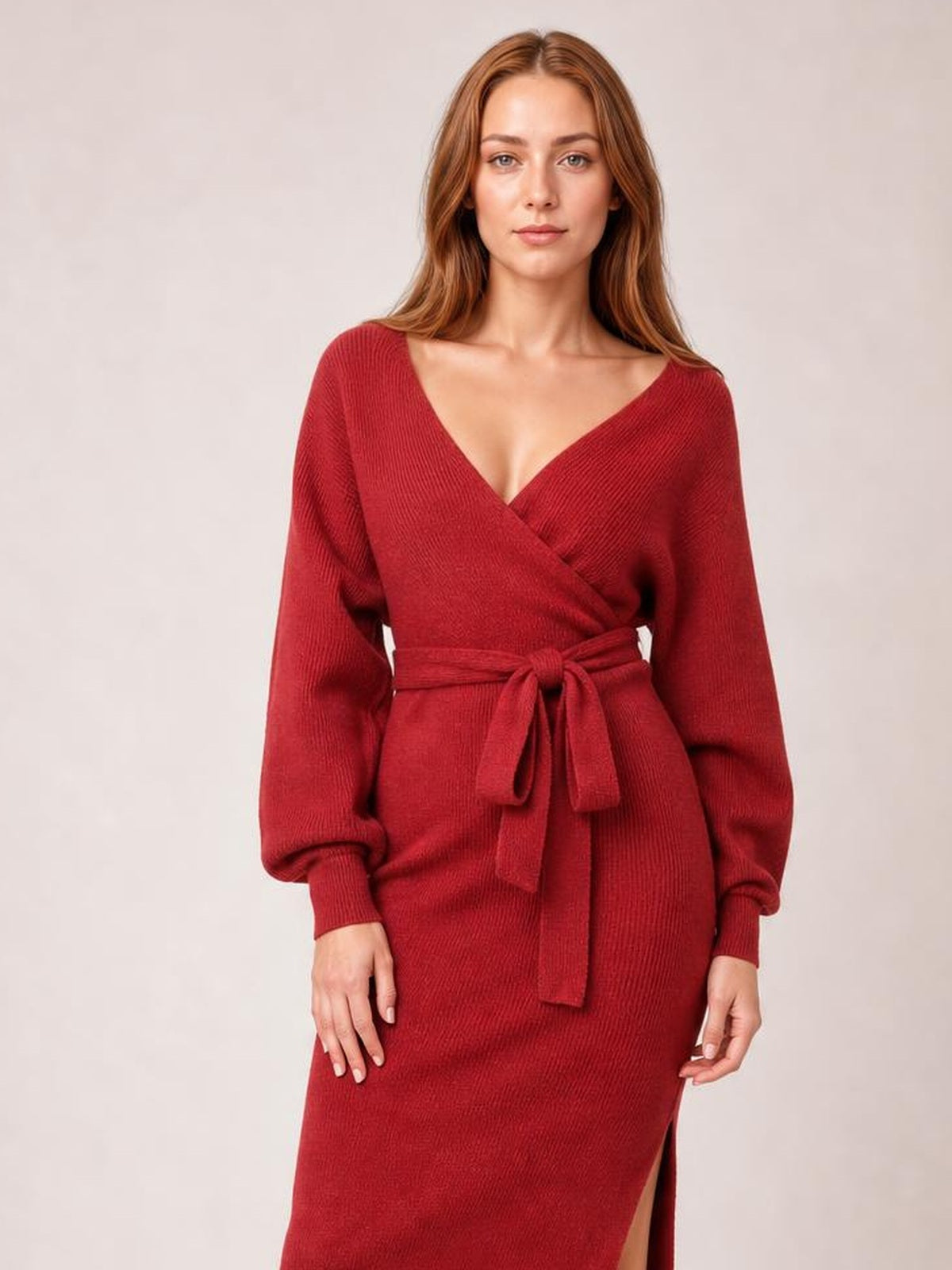 Model wears a red ribbed sweater dress with a surplice neckline, long sleeves, tied bow waist, regular silhouette, and a thigh-high side slit.