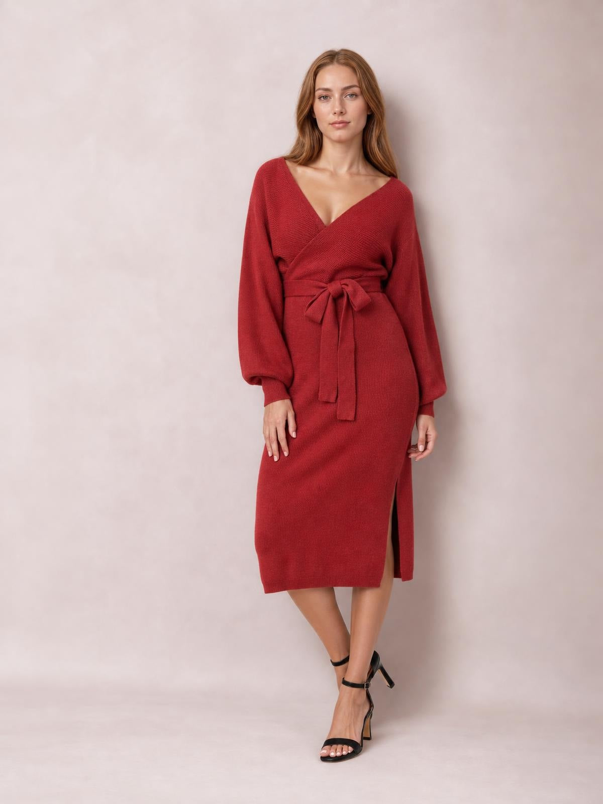 A model poses in a red sweater dress with a surplice neckline, long sleeves, tied bow waist, fitted midi silhouette, and side slit.