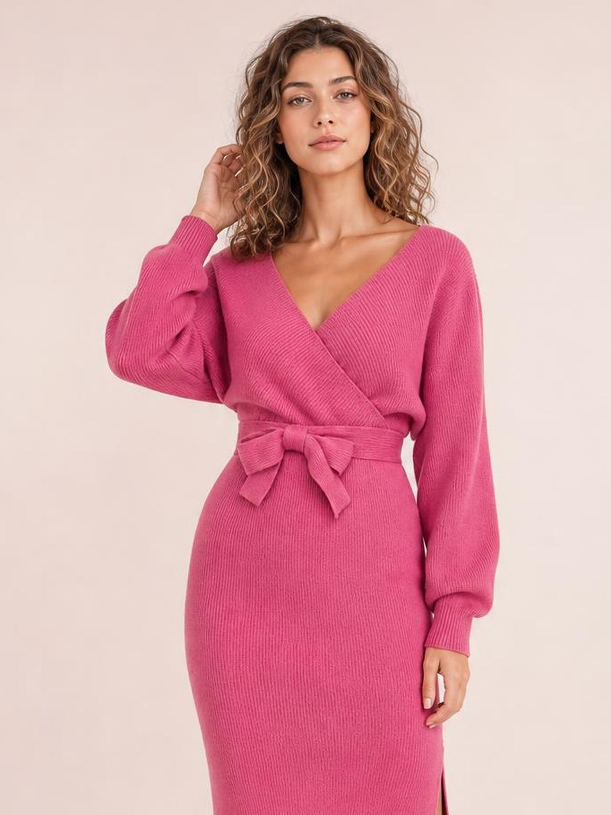 Model poses in a pink ribbed sweater dress with a surplice neckline, long sleeves, tied bow waist, fitted silhouette, and side slit.