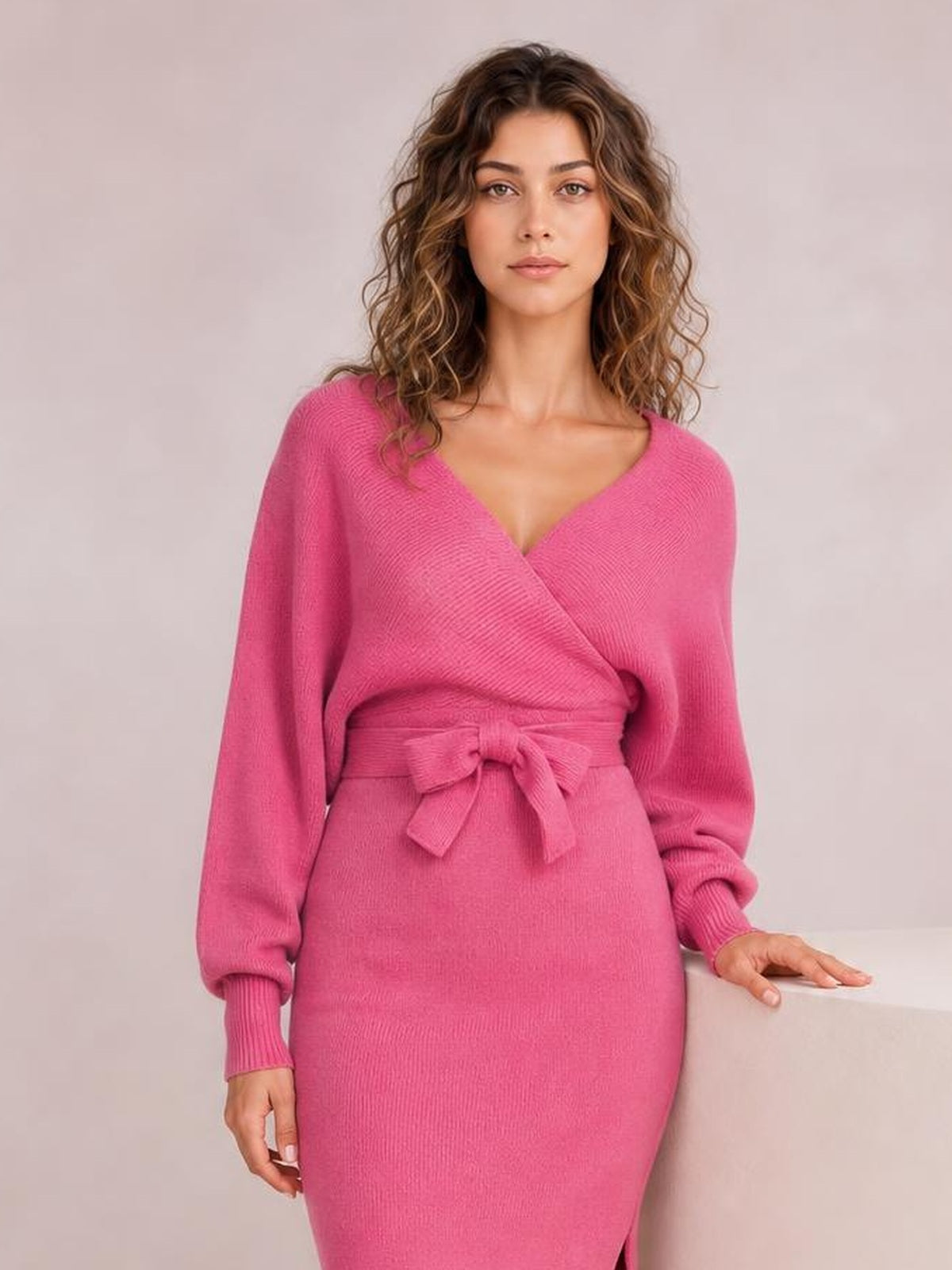 Model in a bright pink sweater dress with surplice neckline, long sleeves, tied bow waist, fitted silhouette, and visible lower-leg slit.