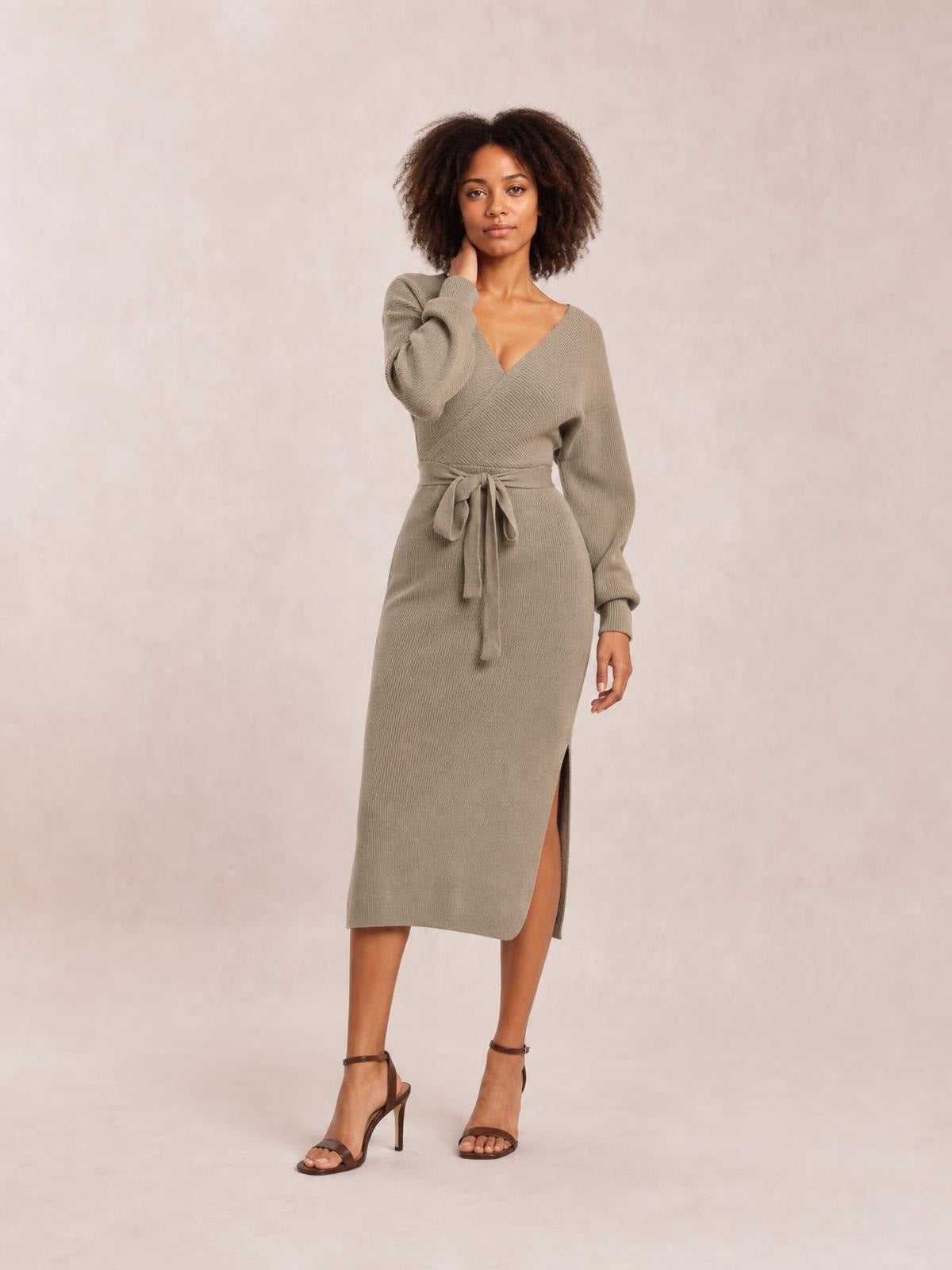 Model poses in a taupe sweater dress with a surplice neckline, long sleeves, tied bow waist, fitted midi silhouette, and side slit.