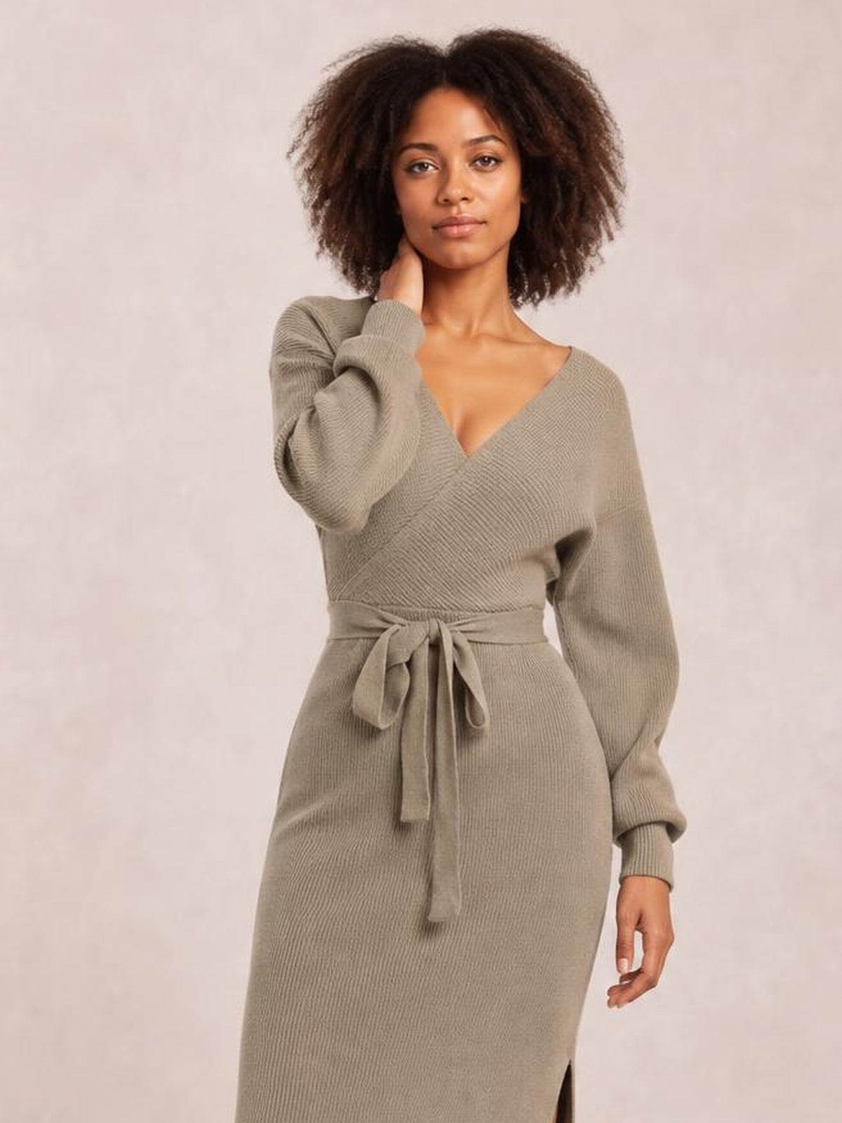 A model poses in a taupe sweater dress with a surplice V-neck, long sleeves, tied bow waist, fitted midi silhouette, and side slit.