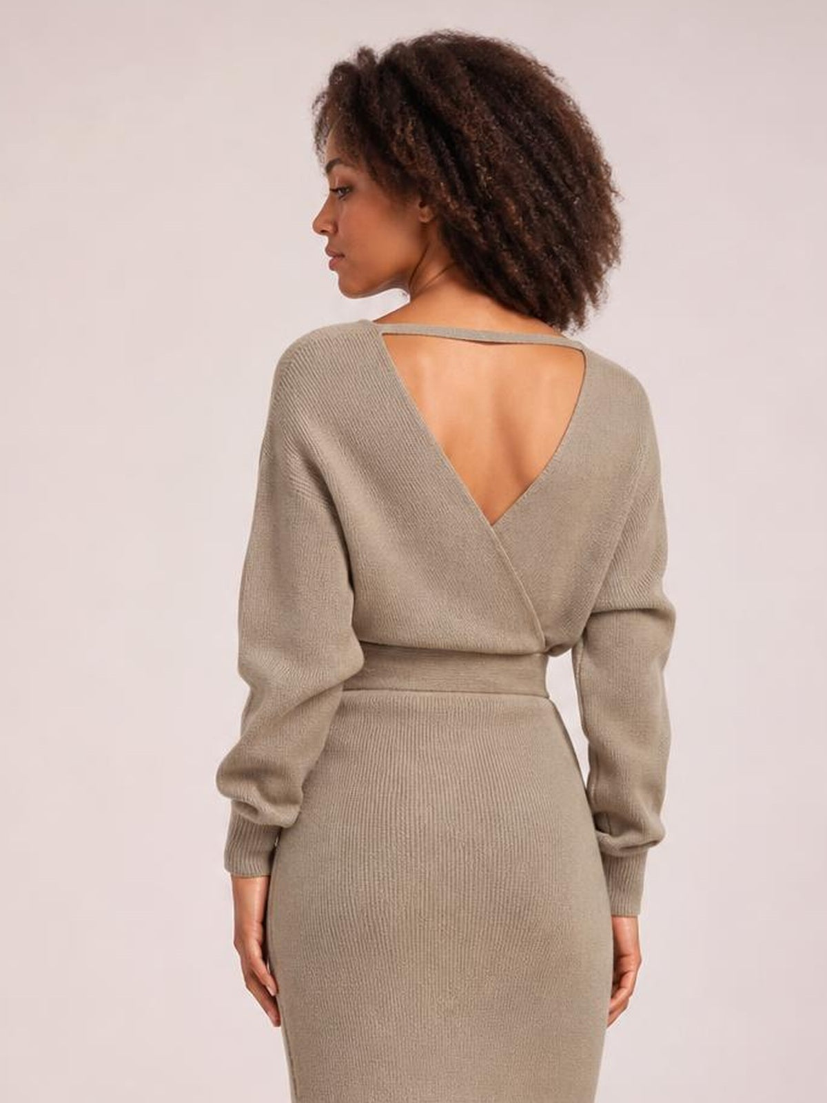 Model shown from behind in a taupe ribbed sweater dress with long sleeves, a crossed surplice back, V-neck opening, defined waist, and fitted midi silhouette.