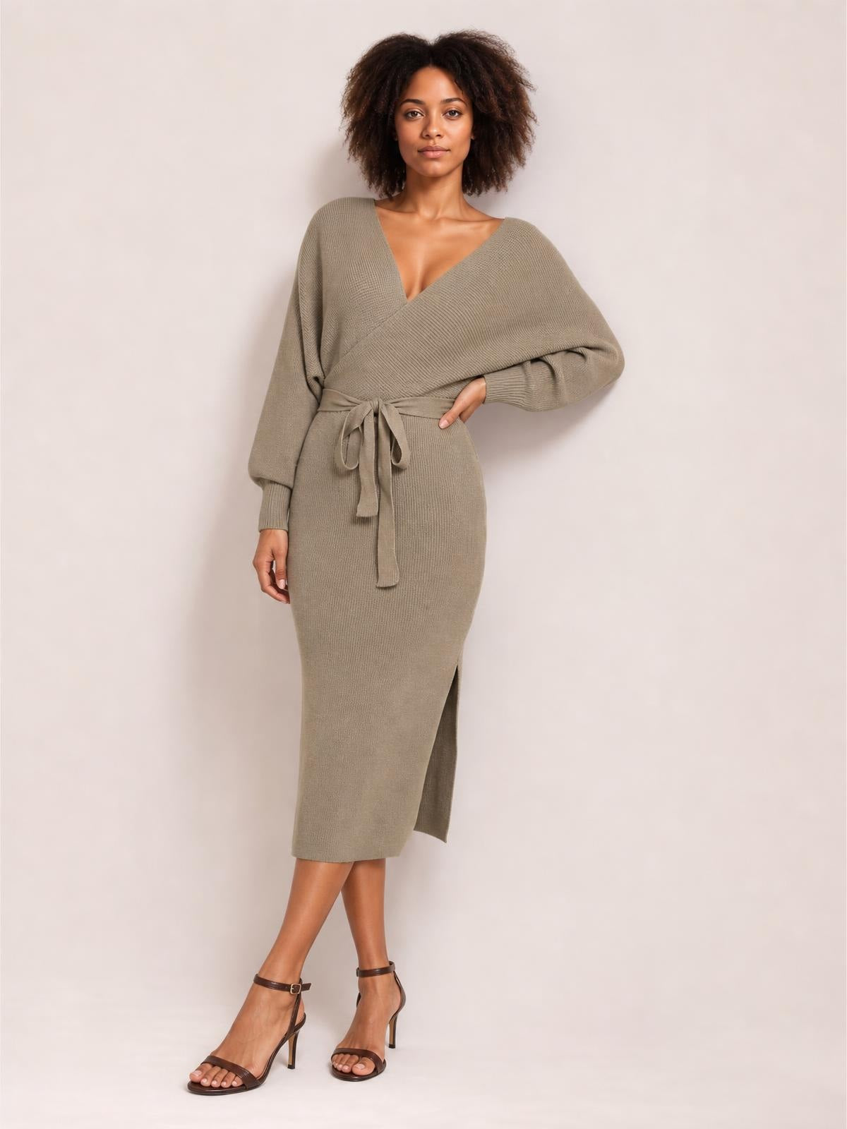 Model poses in a taupe surplice-neck sweater dress with long sleeves, a tied bow waist, fitted midi silhouette, and side slit.
