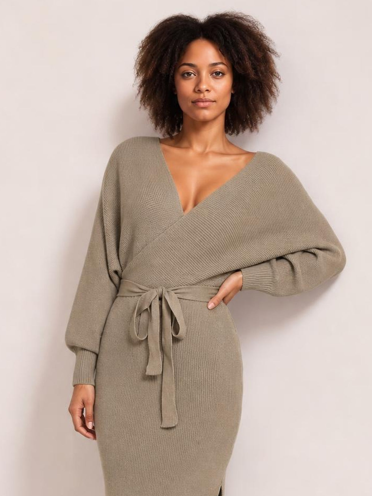 Model poses in a taupe surplice-neck sweater dress with long sleeves, a tied bow waist, ribbed knit texture, fitted silhouette, and side slit.