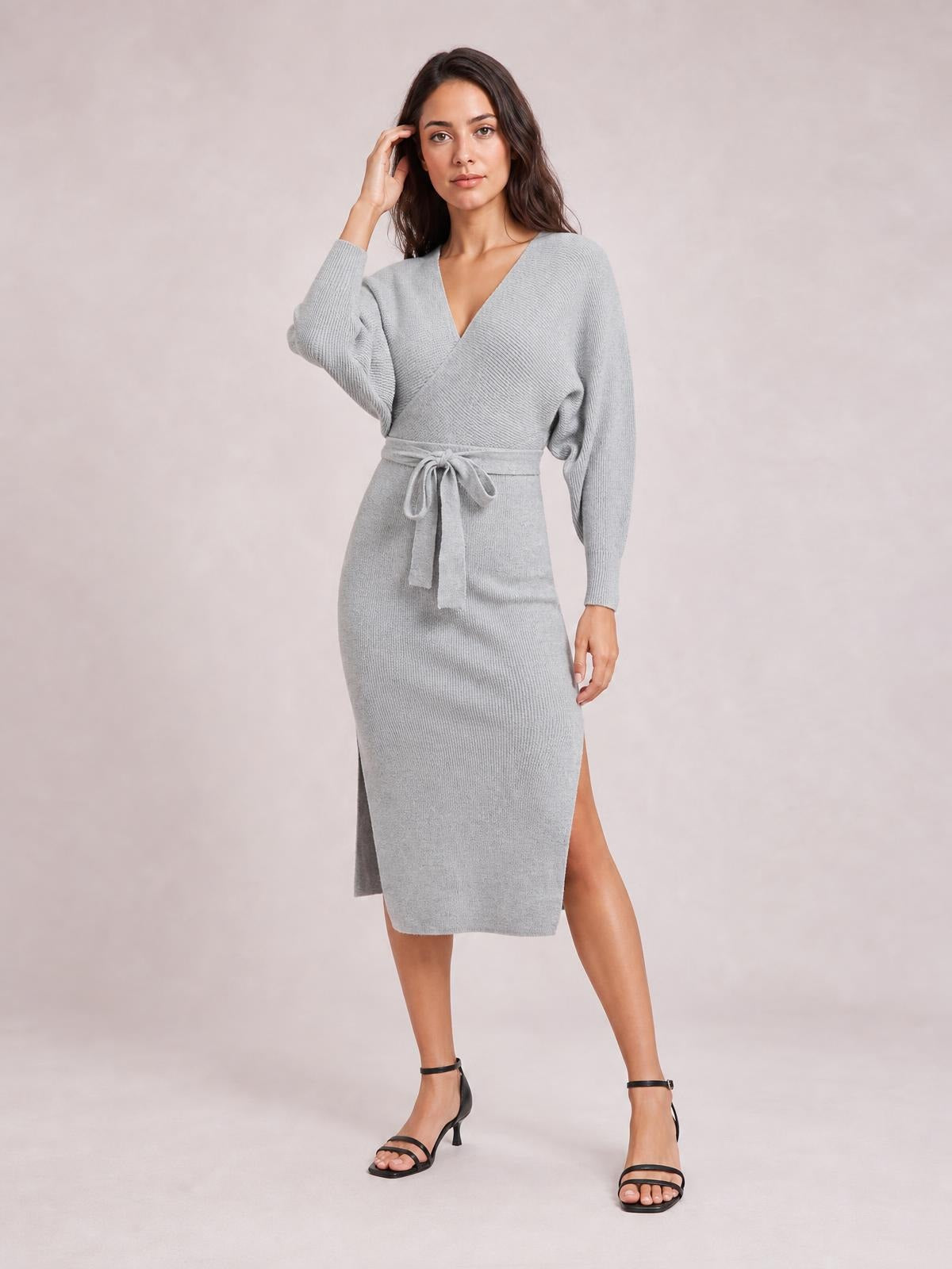 Model poses in a light gray surplice-neck sweater dress with long sleeves, a tied bow waist, fitted midi silhouette, and side slit.