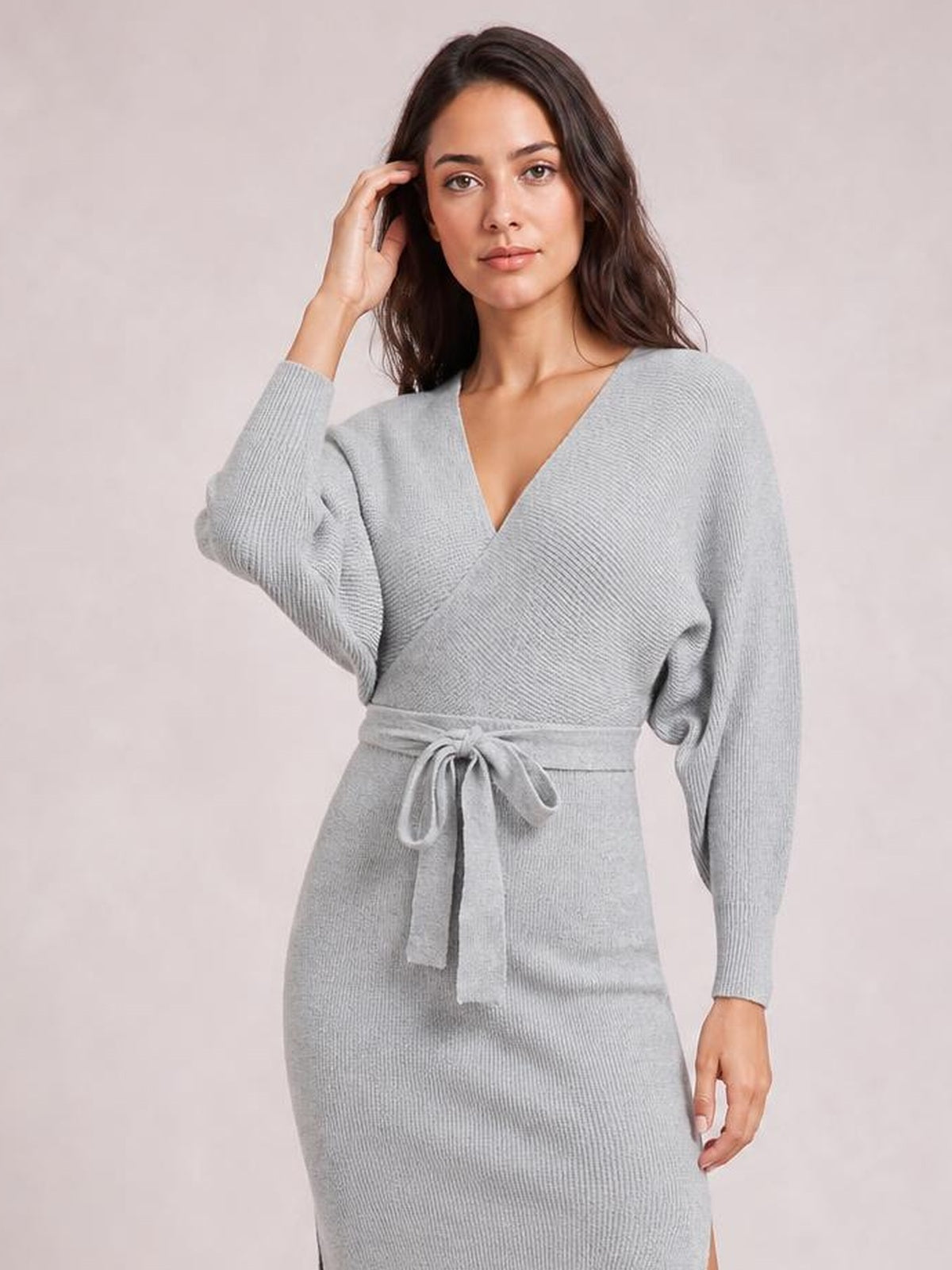 A model poses in a light gray sweater dress with a surplice V-neck, long sleeves, tied bow waist, fitted silhouette, and side slit.