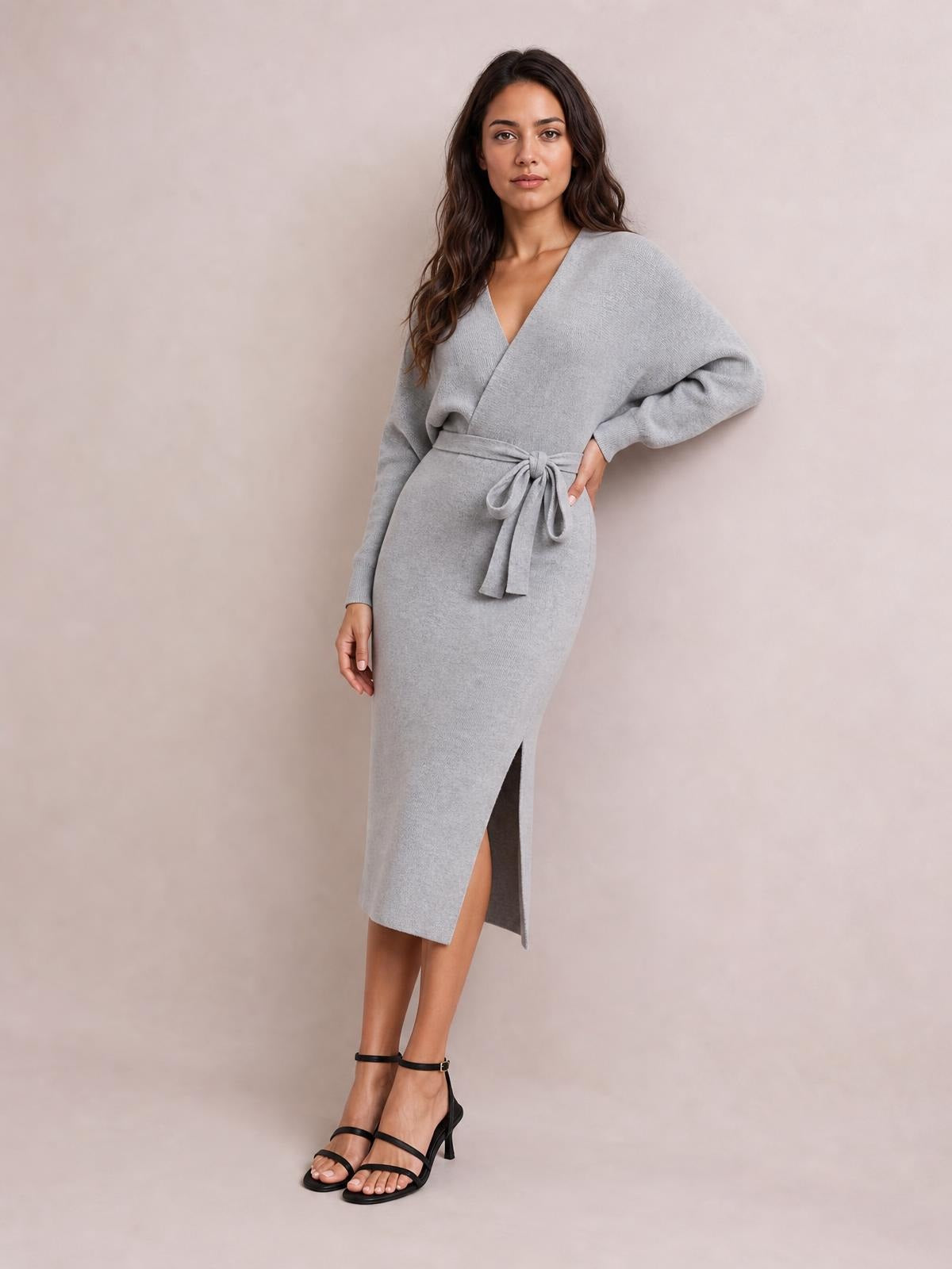 Model poses in a gray surplice neck sweater dress with long sleeves, a tied bow waist, fitted midi silhouette, and side slit.