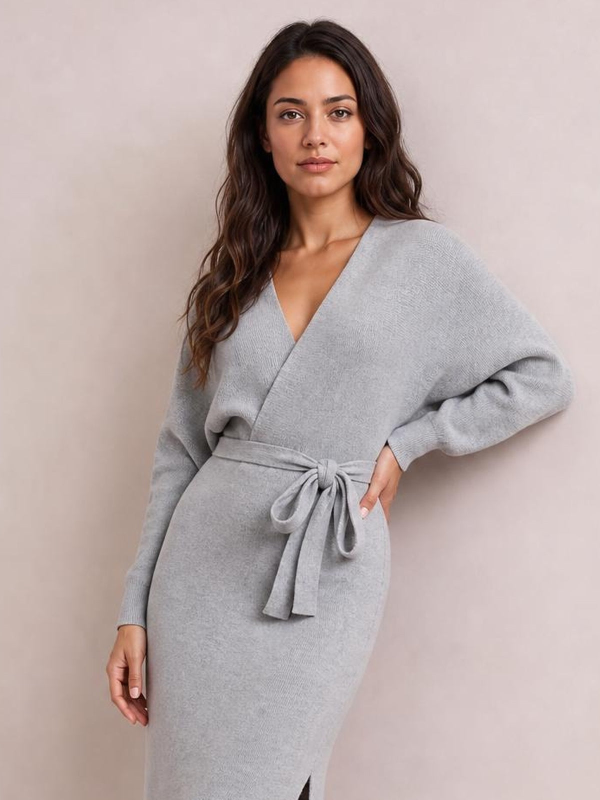 Model wears a light gray surplice-neck sweater dress with long sleeves, a tied bow waist, fitted silhouette, and side slit, posing with one hand on her hip.