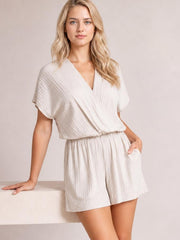 Model in a light beige surplice short sleeve romper with a relaxed silhouette, pleated texture, elastic waist, and roomy shorts, posing with one hand in a pocket.