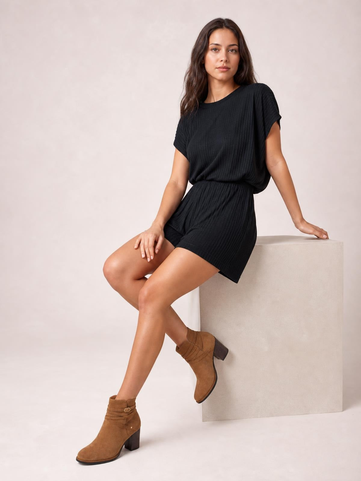 A model sits with crossed legs wearing a black short-sleeve romper with a relaxed top, ribbed texture, elastic waist, and loose shorts.