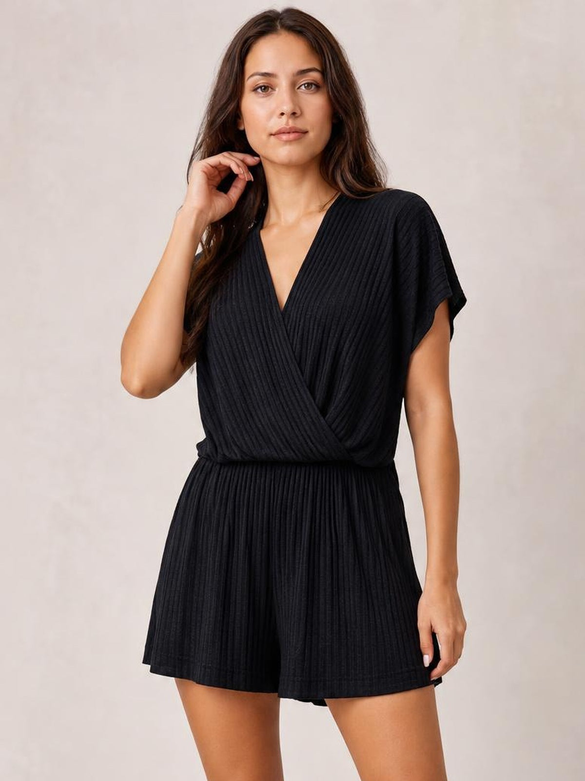 A model poses in a black surplice short-sleeve romper with a V-neck wrap front, vertical ribbed texture, defined waist, and relaxed shorts.