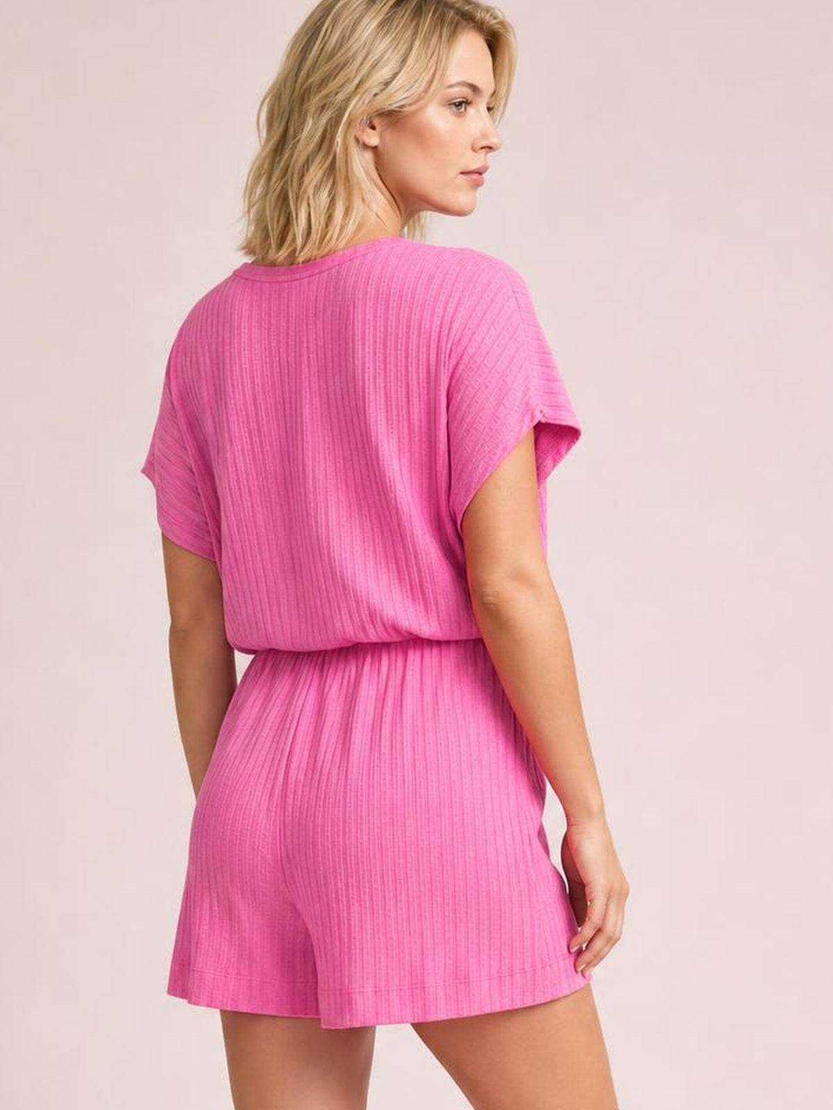 Model shown from behind wearing a bright pink short-sleeve romper with a softly textured finish, relaxed silhouette, defined waist, and flared shorts.