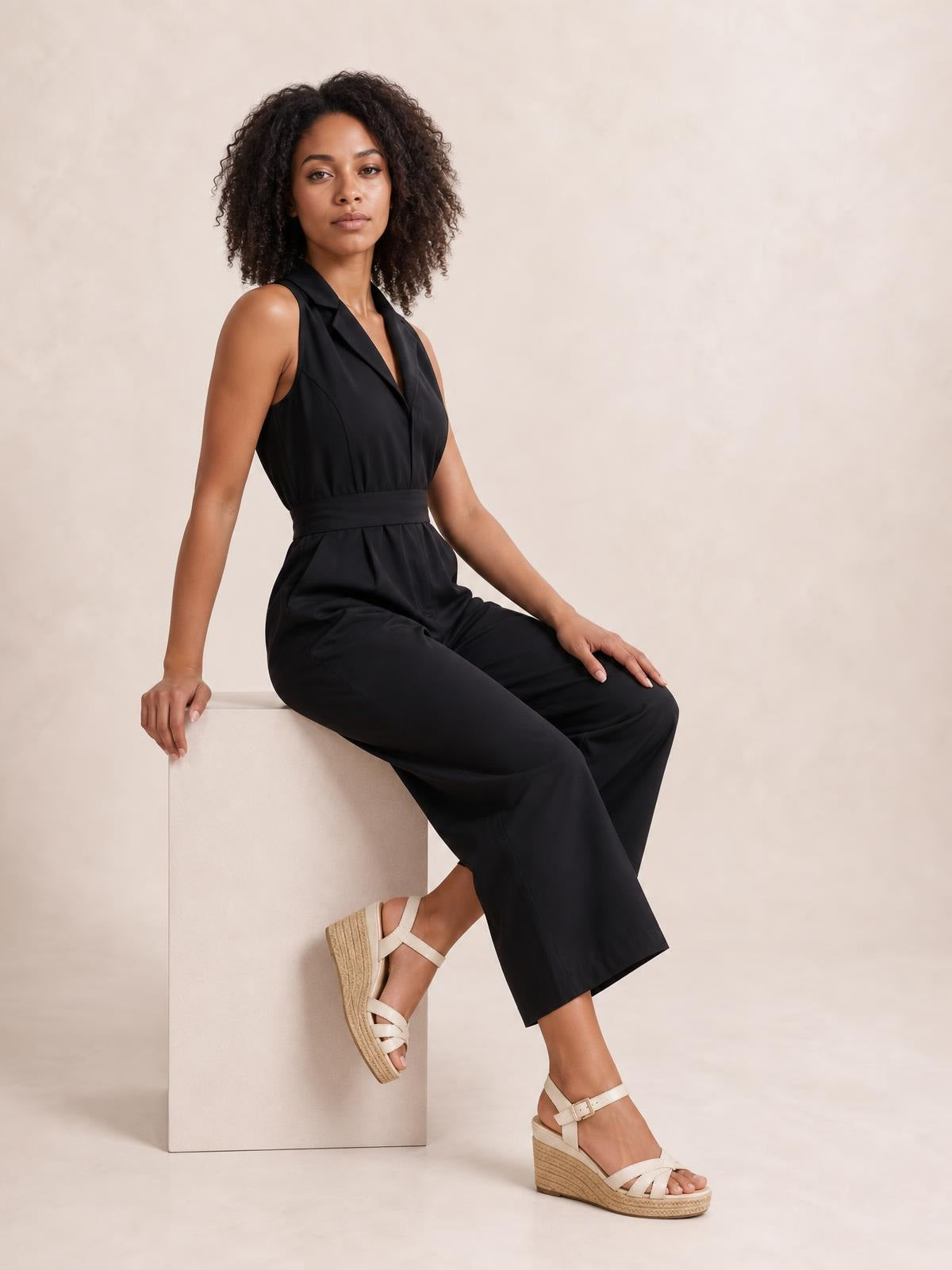 A model sits wearing a black sleeveless wide-leg jumpsuit with a surplice neckline, defined tied waist, and relaxed flowing silhouette.