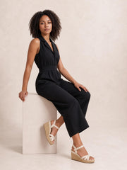A model sits wearing a black sleeveless wide-leg jumpsuit with a surplice neckline, defined tied waist, and relaxed flowing silhouette.