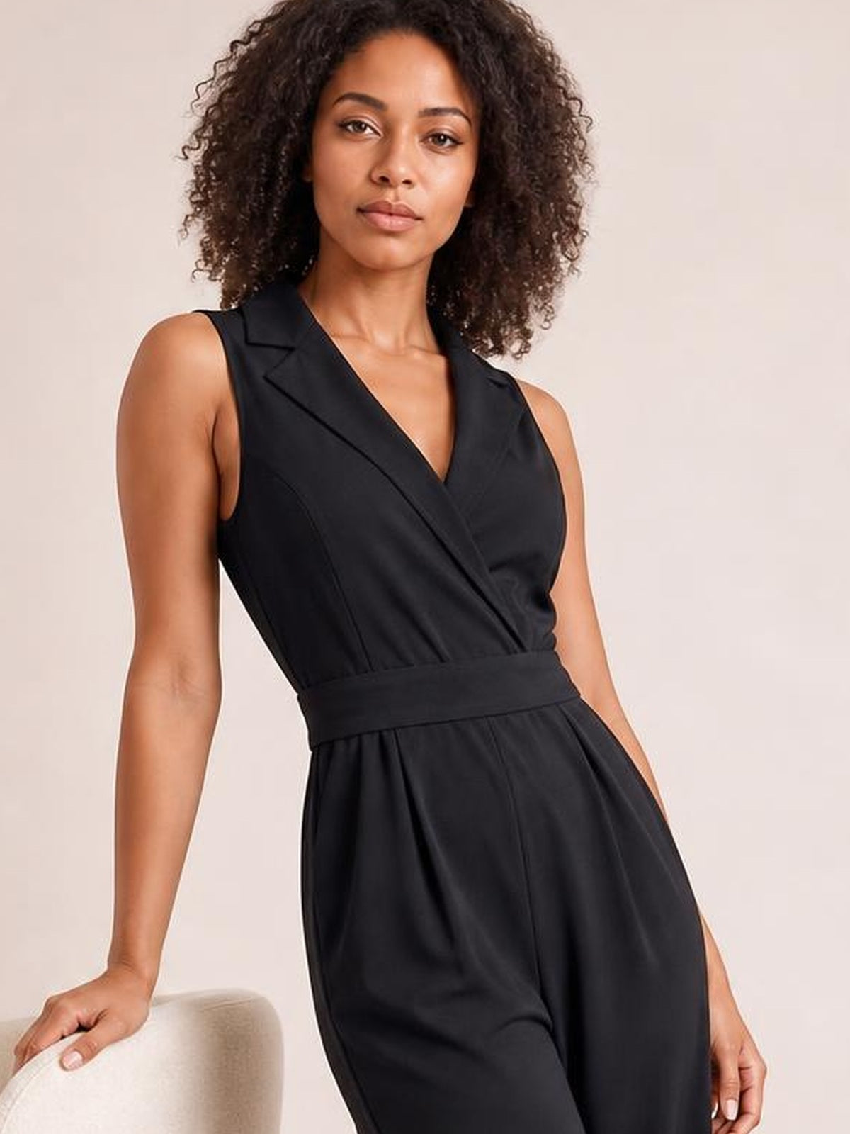 A model poses in a black sleeveless surplice jumpsuit with a defined waist, wide-leg silhouette, tailored lapels, and a flowing drape.