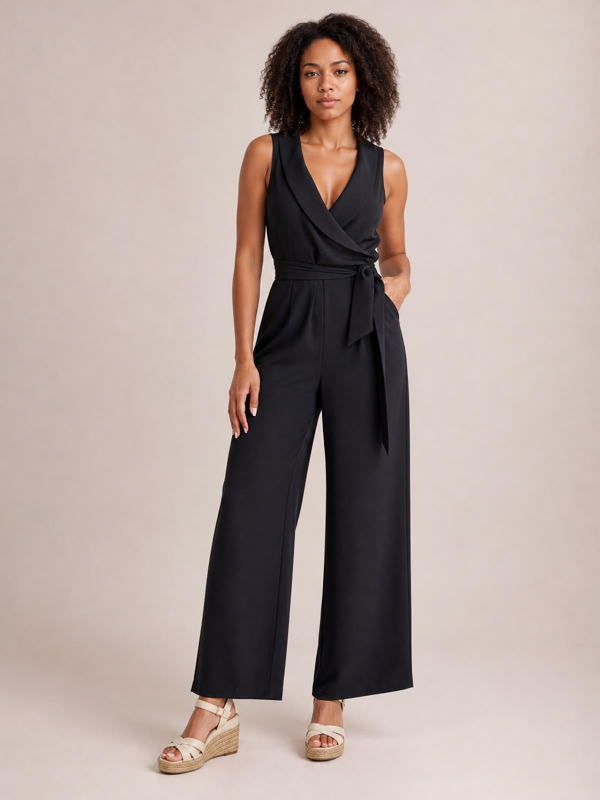 Model wears a black surplice sleeveless jumpsuit with a tied waist, pocketed design, and flowing wide-leg silhouette.