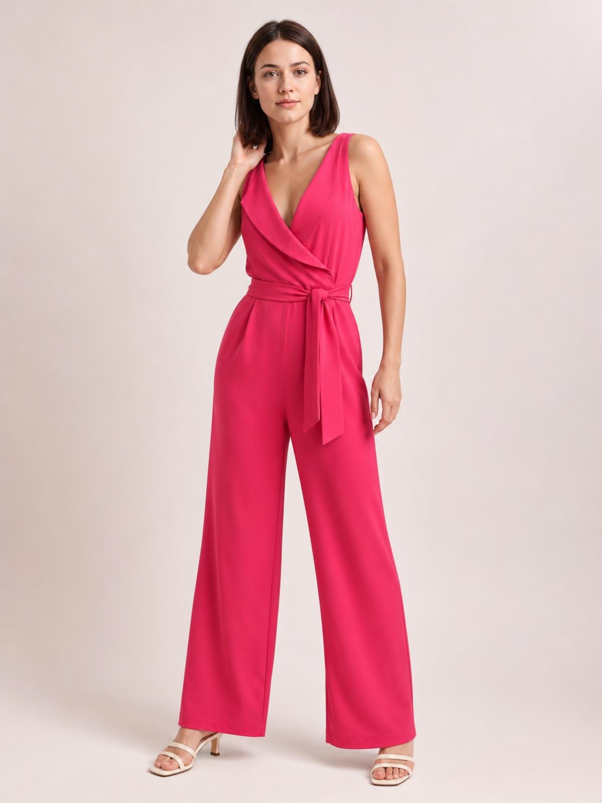 Model posing in a bright pink surplice sleeveless jumpsuit with a tied waist, flowing wide-leg silhouette, and visible side pockets.