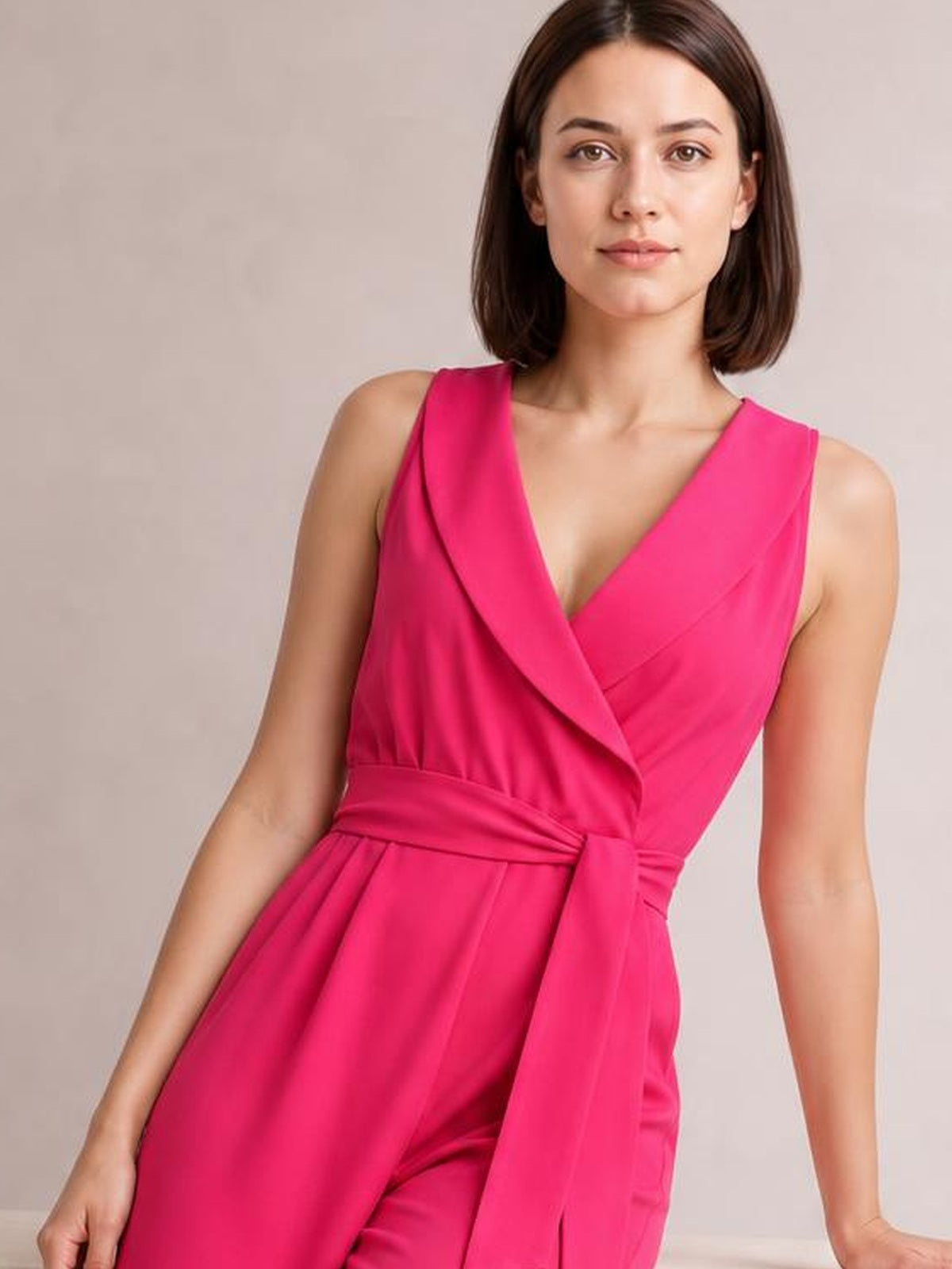 Model poses in a bright pink sleeveless jumpsuit with a surplice neckline, tied waist, and flowing wide-leg silhouette.