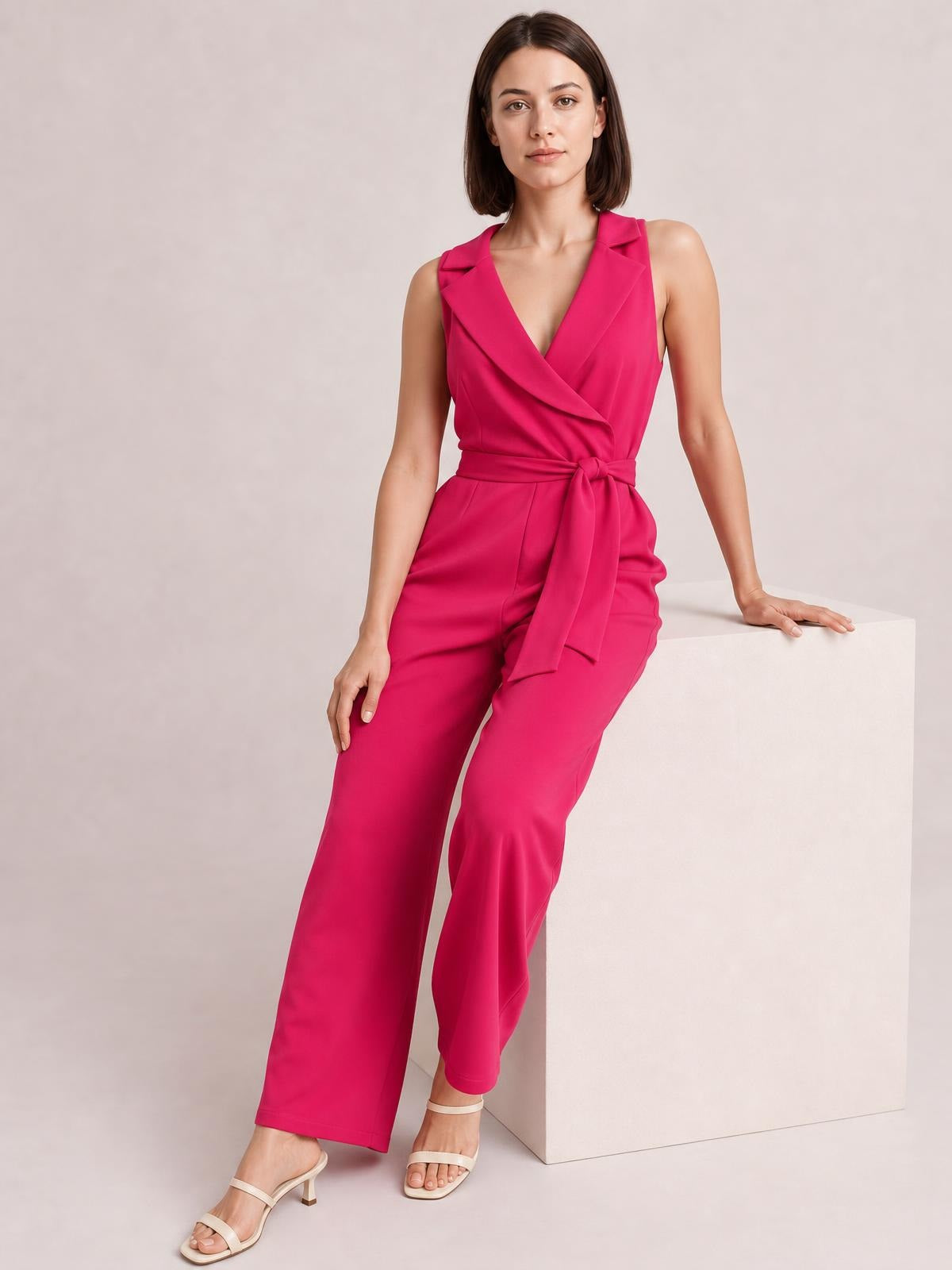 A model poses in a bright pink surplice sleeveless jumpsuit with a wrap-style neckline, tied waist, and flowing wide-leg silhouette.