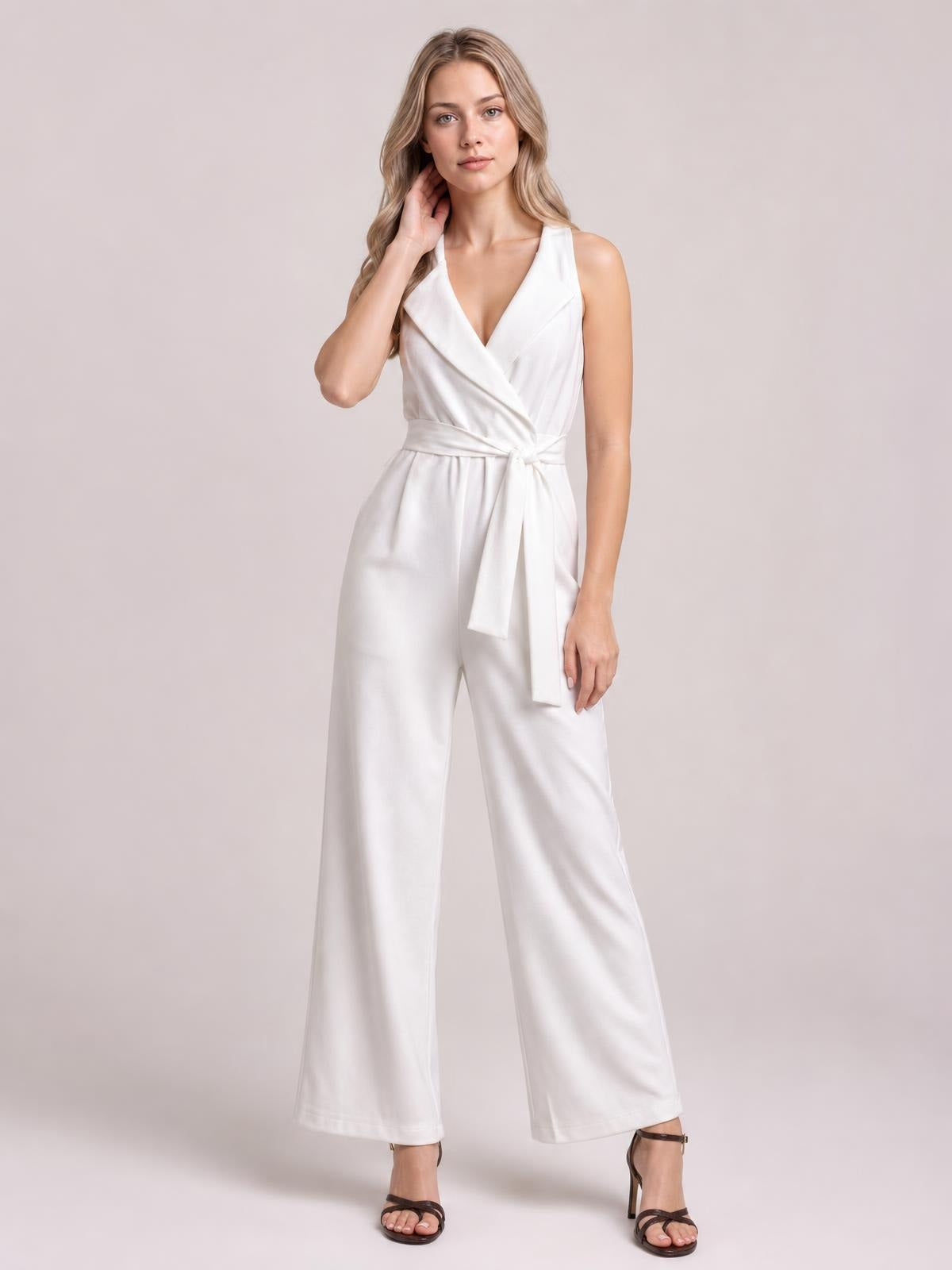Model poses in a white sleeveless surplice jumpsuit with a tied waist, wide-leg silhouette, wrap-style bodice, and side pockets.