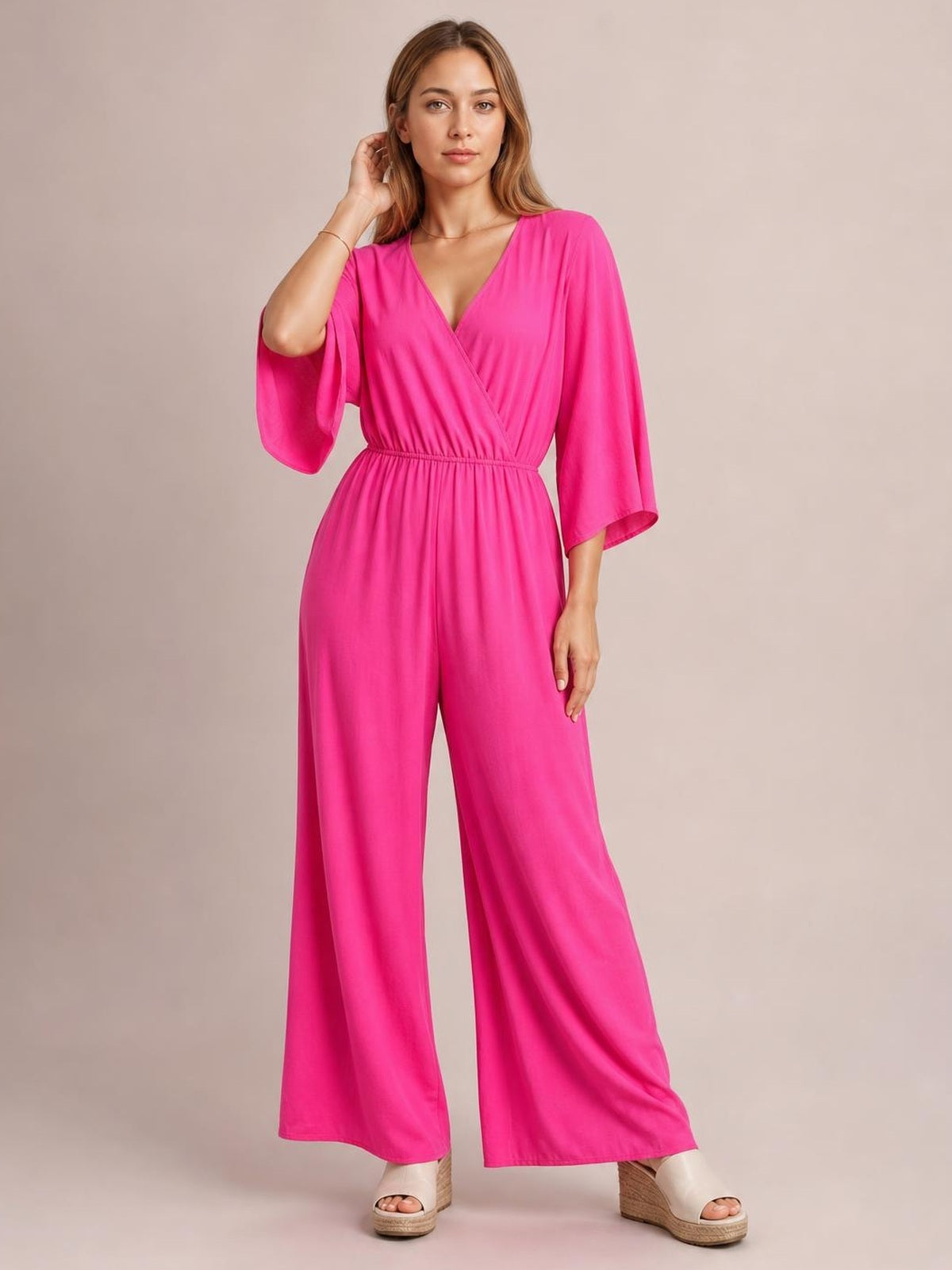 Model poses in a bright pink surplice jumpsuit with ruched waist, wide legs, three-quarter sleeves, and a flattering full-length silhouette.