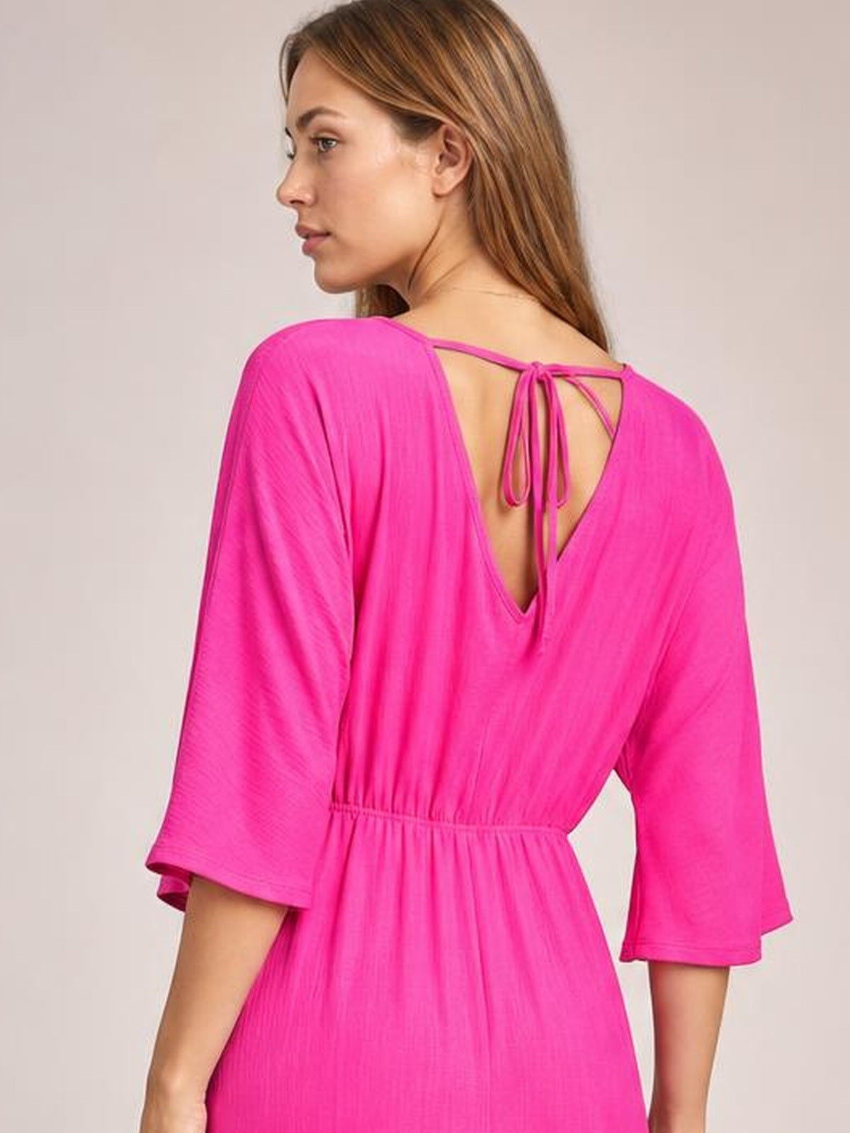 Model turns to show a hot pink jumpsuit with a ruched waist, three-quarter sleeves, deep V-back, and tied open-back detail.