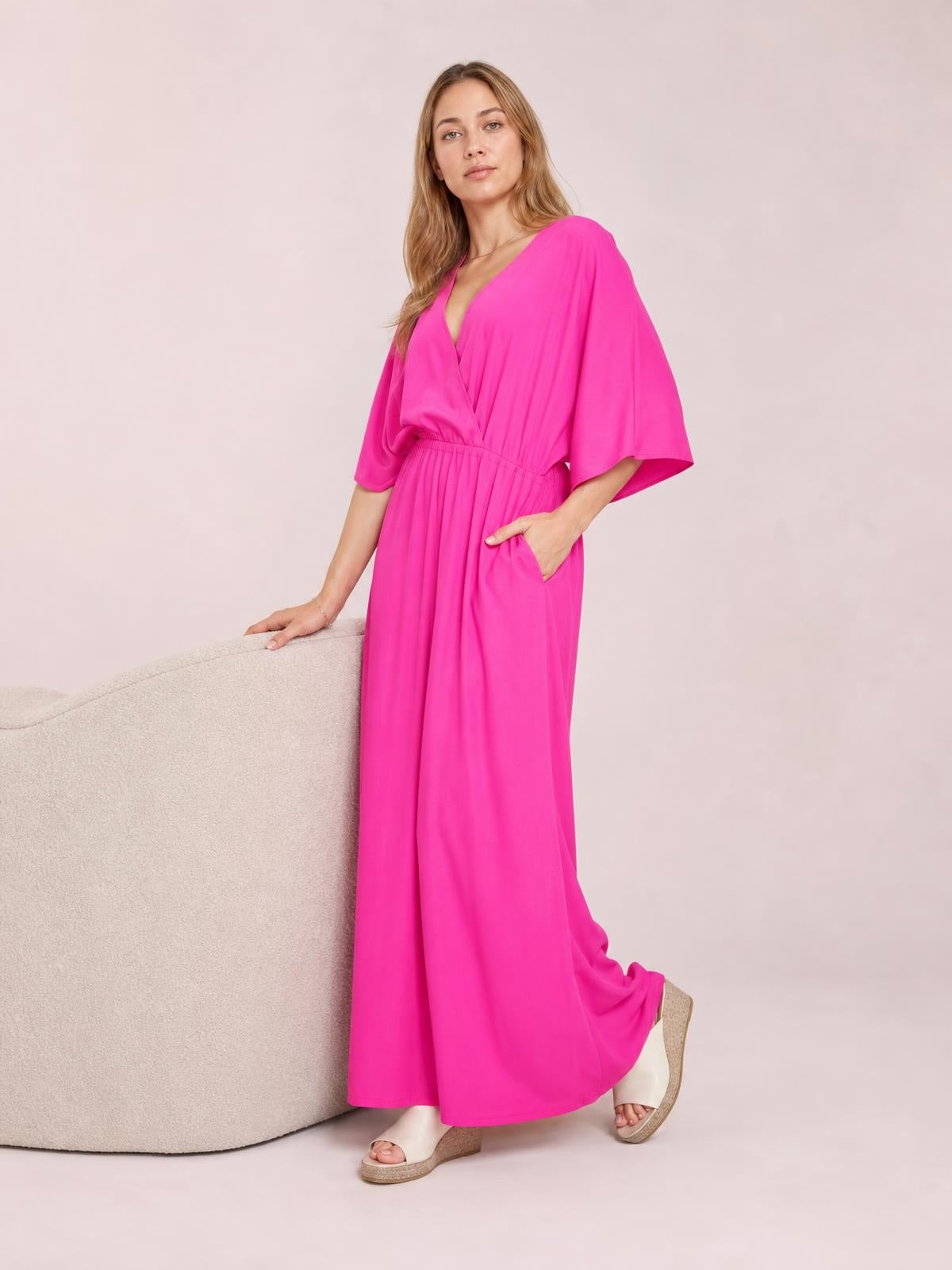 Model poses in a bright pink surplice wide-leg jumpsuit with a ruched waist, tied details, flowing silhouette, and visible side pockets.