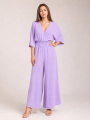 Model poses in a lavender surplice jumpsuit with a ruched waist, tied wrap-style bodice, flowing wide legs, and practical pockets.