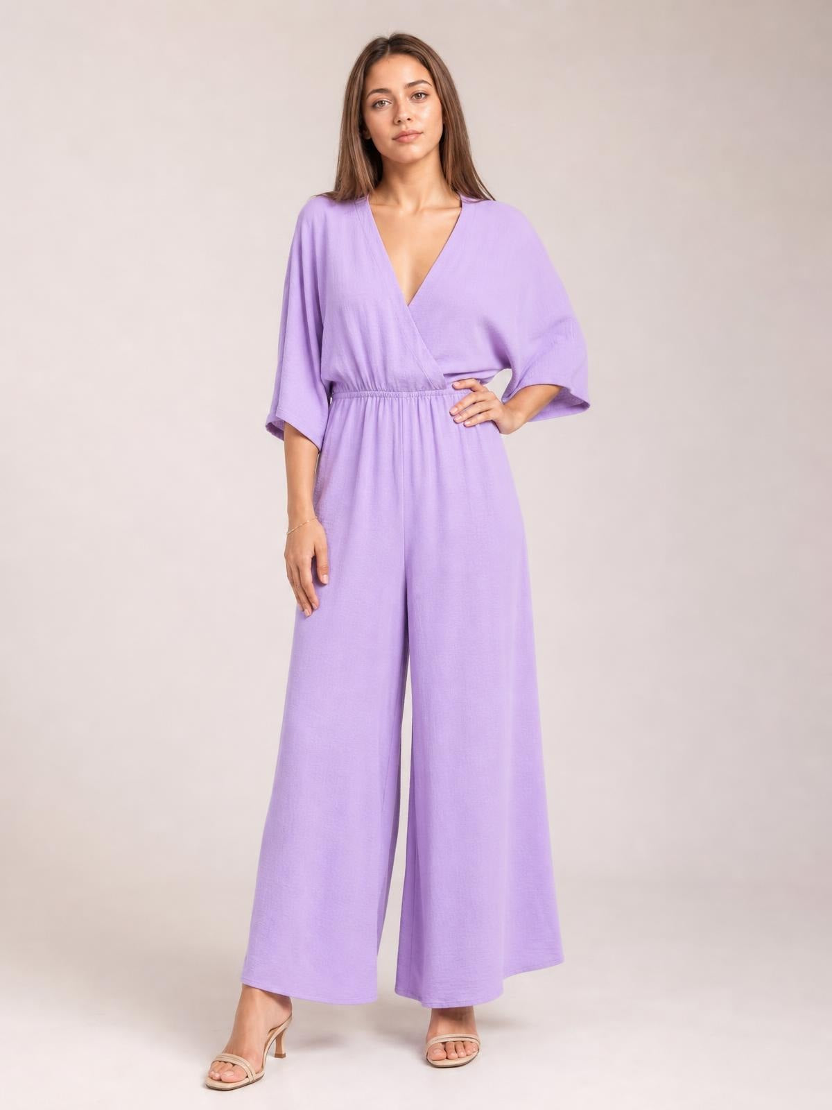 Model poses in a lavender surplice jumpsuit with a ruched waist, tied wrap-style bodice, flowing wide legs, and practical pockets.