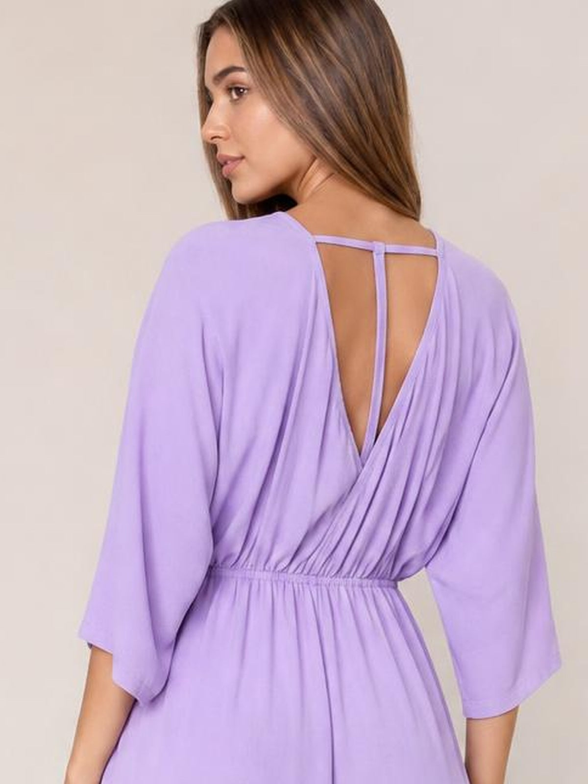 A model poses turned away in a lavender surplice jumpsuit with a ruched elastic waist, open V-back with straps, and relaxed three-quarter sleeves.