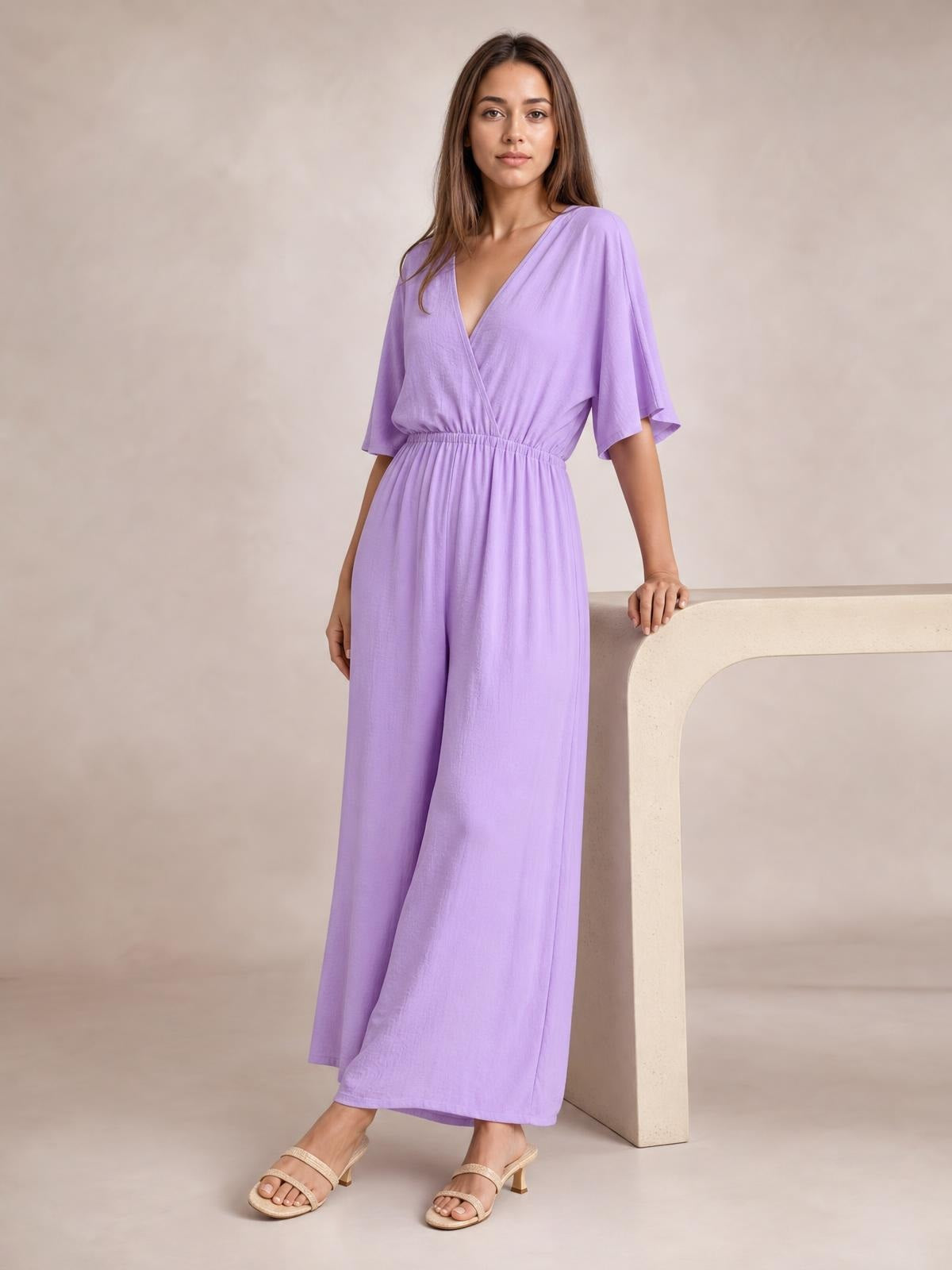 A model wears a lavender surplice jumpsuit with flutter sleeves, a ruched waist, tied detail, and flowing wide legs while resting one hand on a console.