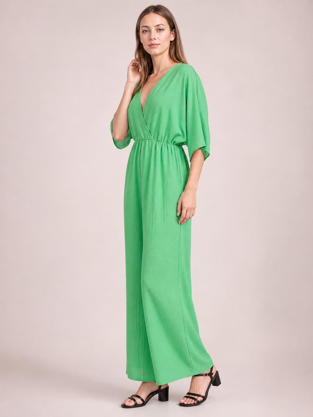 Model poses in a green surplice jumpsuit with a ruched waist, tied details, flutter sleeves, pockets, and flowing wide legs.