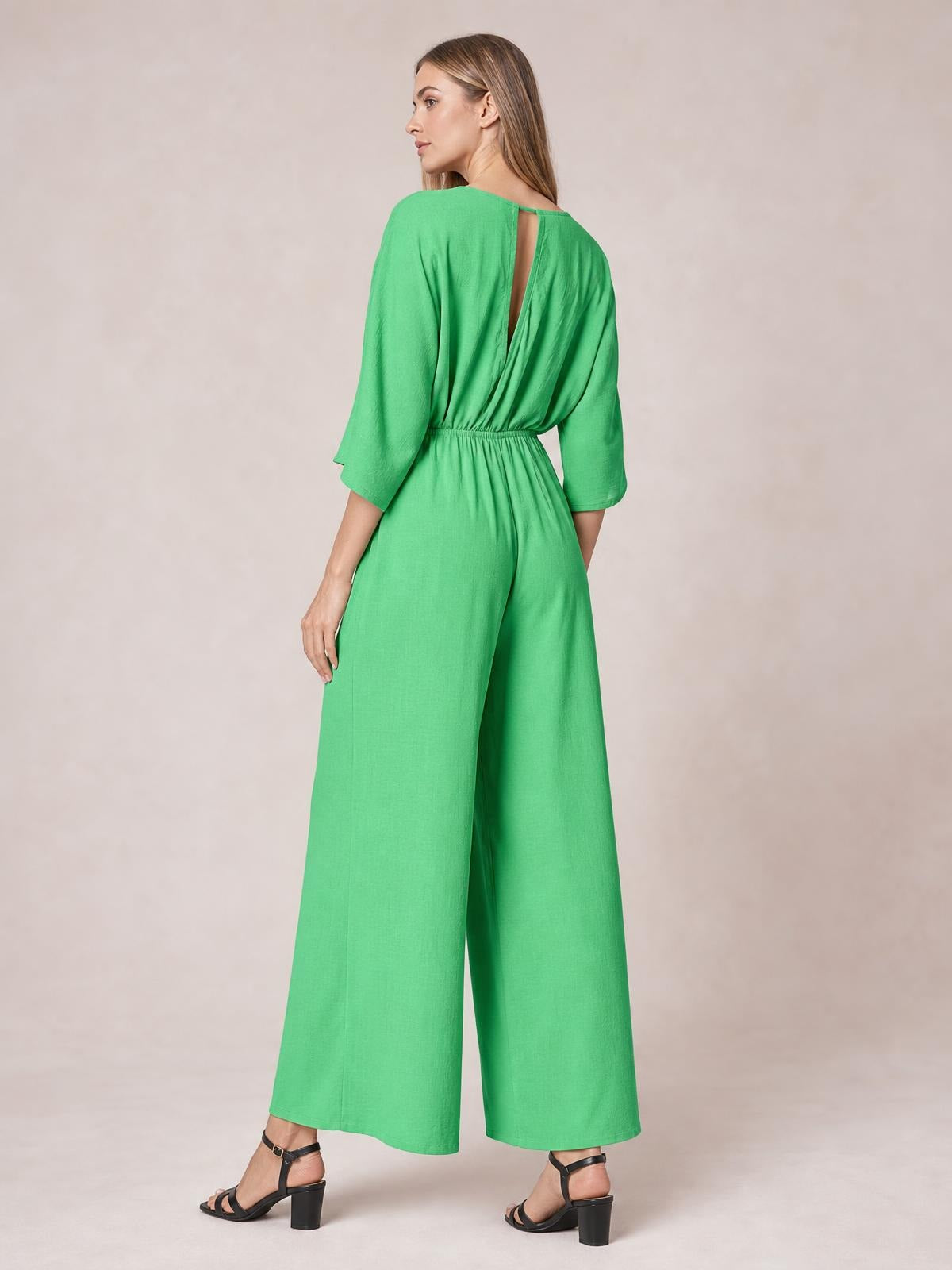 A model poses from behind in a bright green wide-leg jumpsuit with three-quarter sleeves, a ruched elastic waist, and an open back detail.