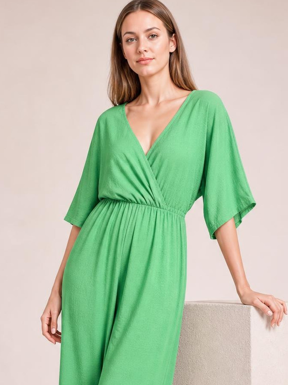 Model wears a green surplice jumpsuit with a ruched waist, tied wrap-style bodice, wide-leg silhouette, and three-quarter sleeves.