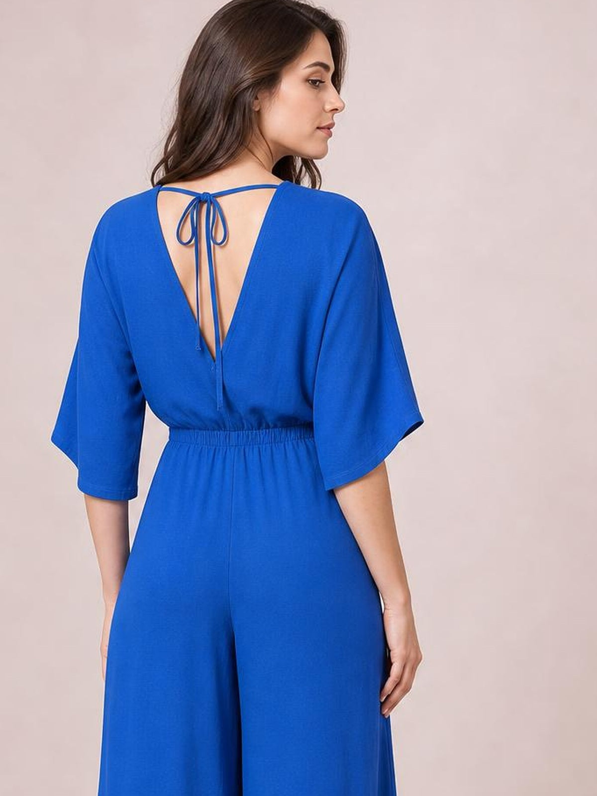 A model stands turned away in a royal blue wide-leg jumpsuit with a ruched elastic waist, wide sleeves, tied open V-back, and relaxed silhouette.