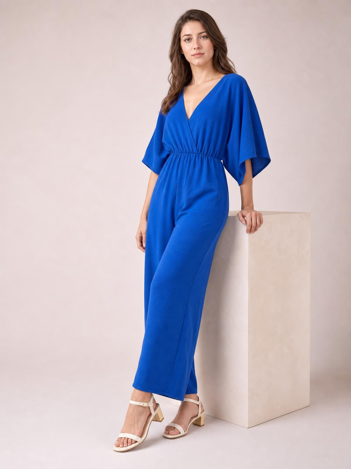 Model leans against a block wearing a bright blue surplice jumpsuit with wide legs, ruched waist, and flowing batwing sleeves.