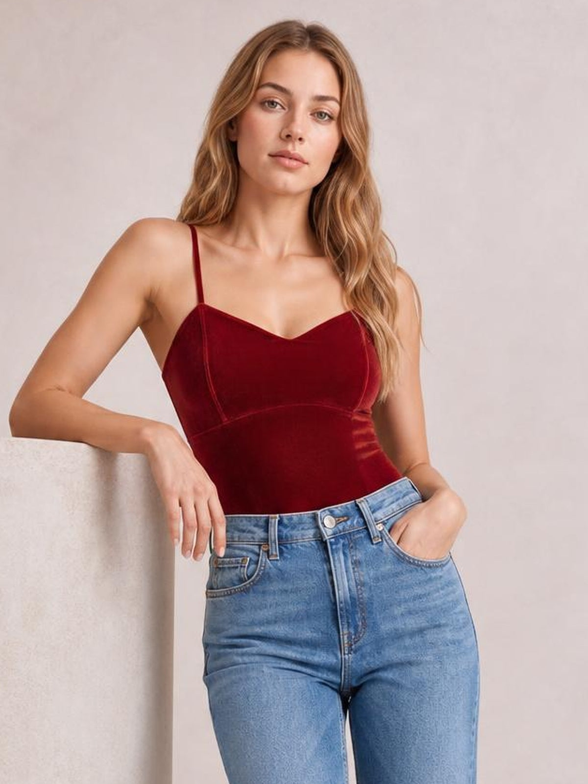 Model wears a burgundy sweetheart neck cropped cami with thin straps and a fitted silhouette, paired with jeans while leaning casually against a ledge.